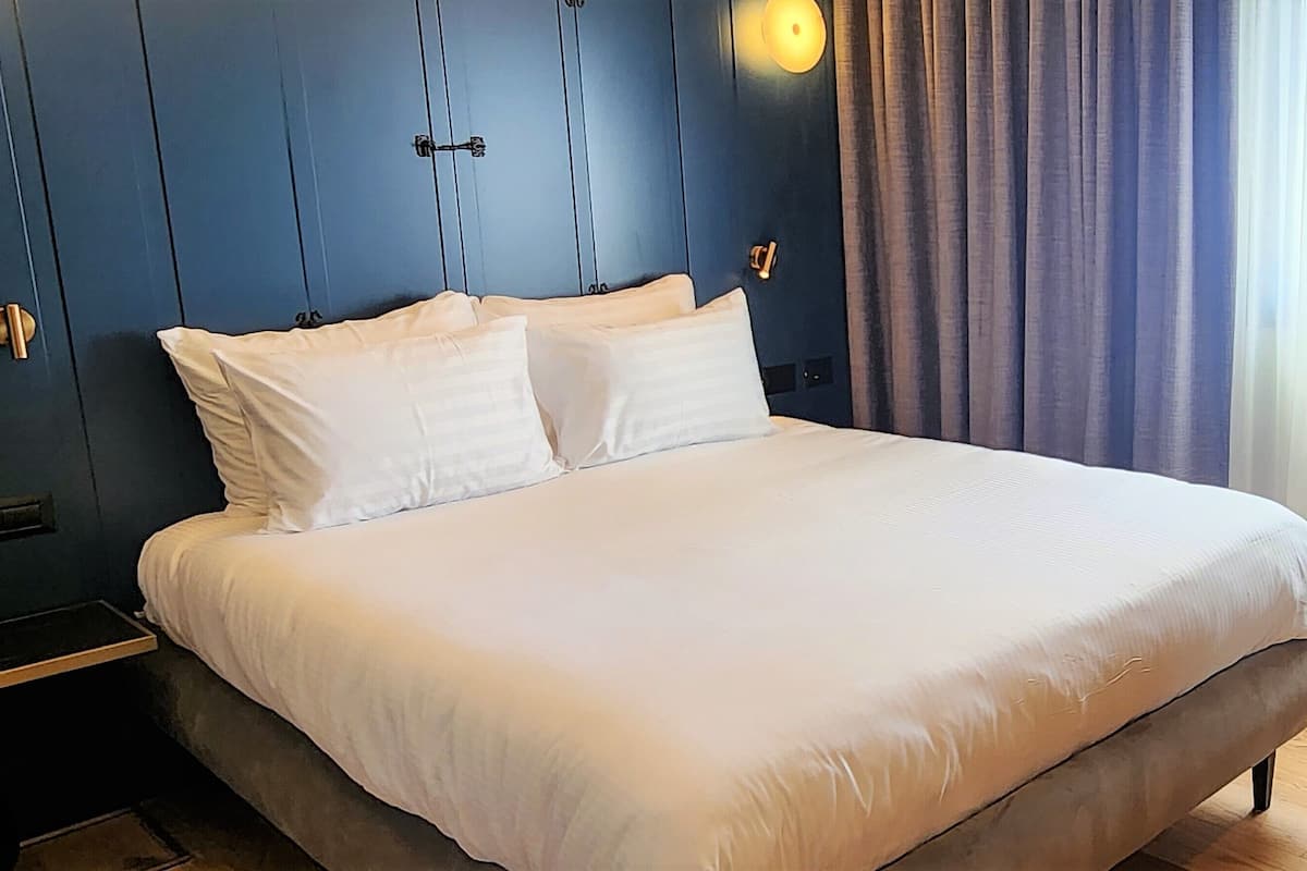 A neatly made bed with white linens, a dark blue headboard, and a window with curtains.