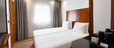 Una habitación de hotel con cama doble, cabecero de madera, escritorio con cafetera y ventana con cortinas.