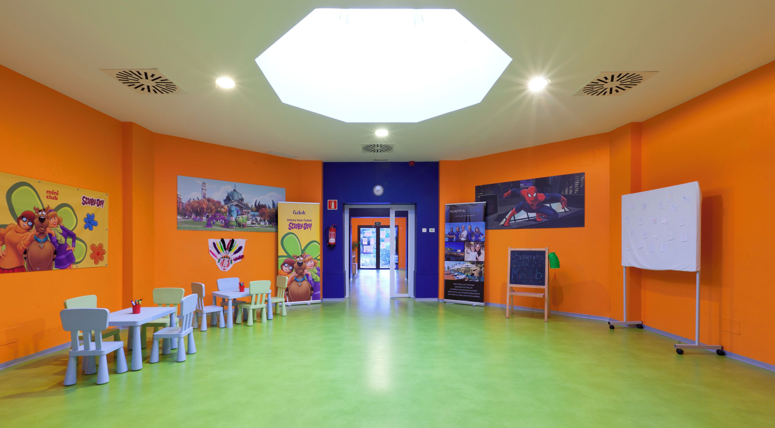 Une pièce aux couleurs vives, avec des murs orange, un sol vert et une grande lucarne octogonale. On y trouve des tables et des chaises disposées pour accueillir les visiteurs, ainsi que divers affiches et œuvres d’art accrochées aux murs.