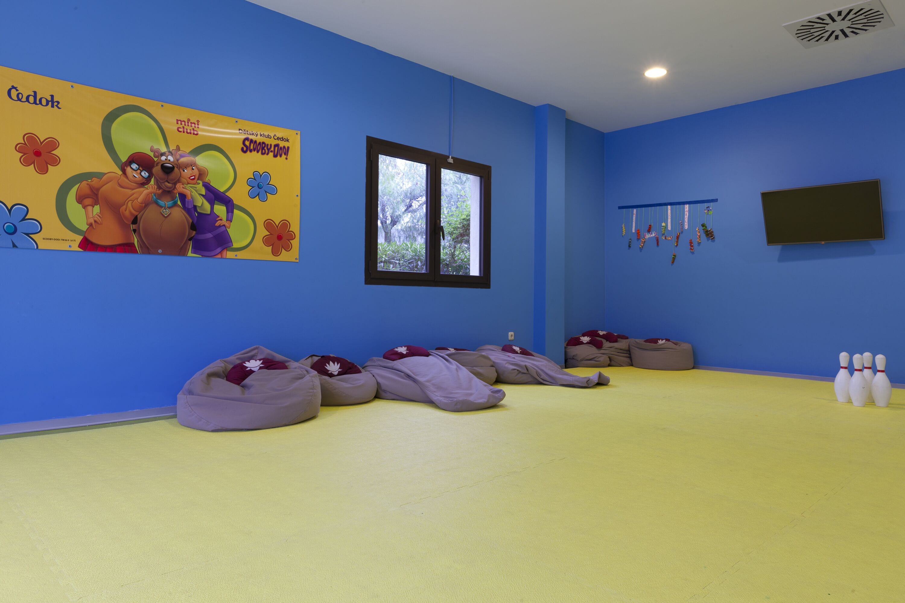 Una habitación con paredes azules, un ventanal grande, pufs, televisor y una zona de bolos.