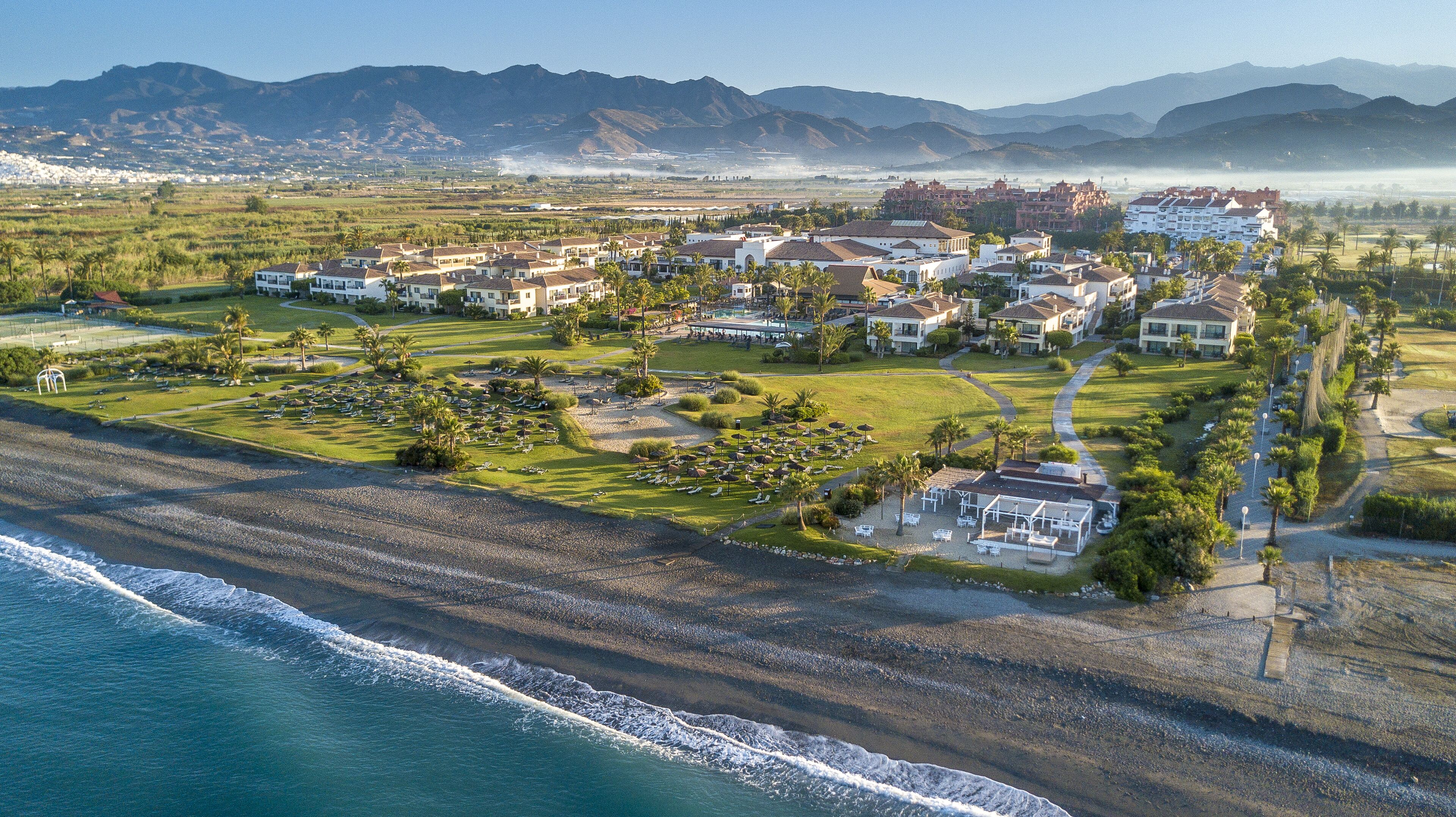 Un complejo turístico frente al mar con varias construcciones y montañas al fondo.