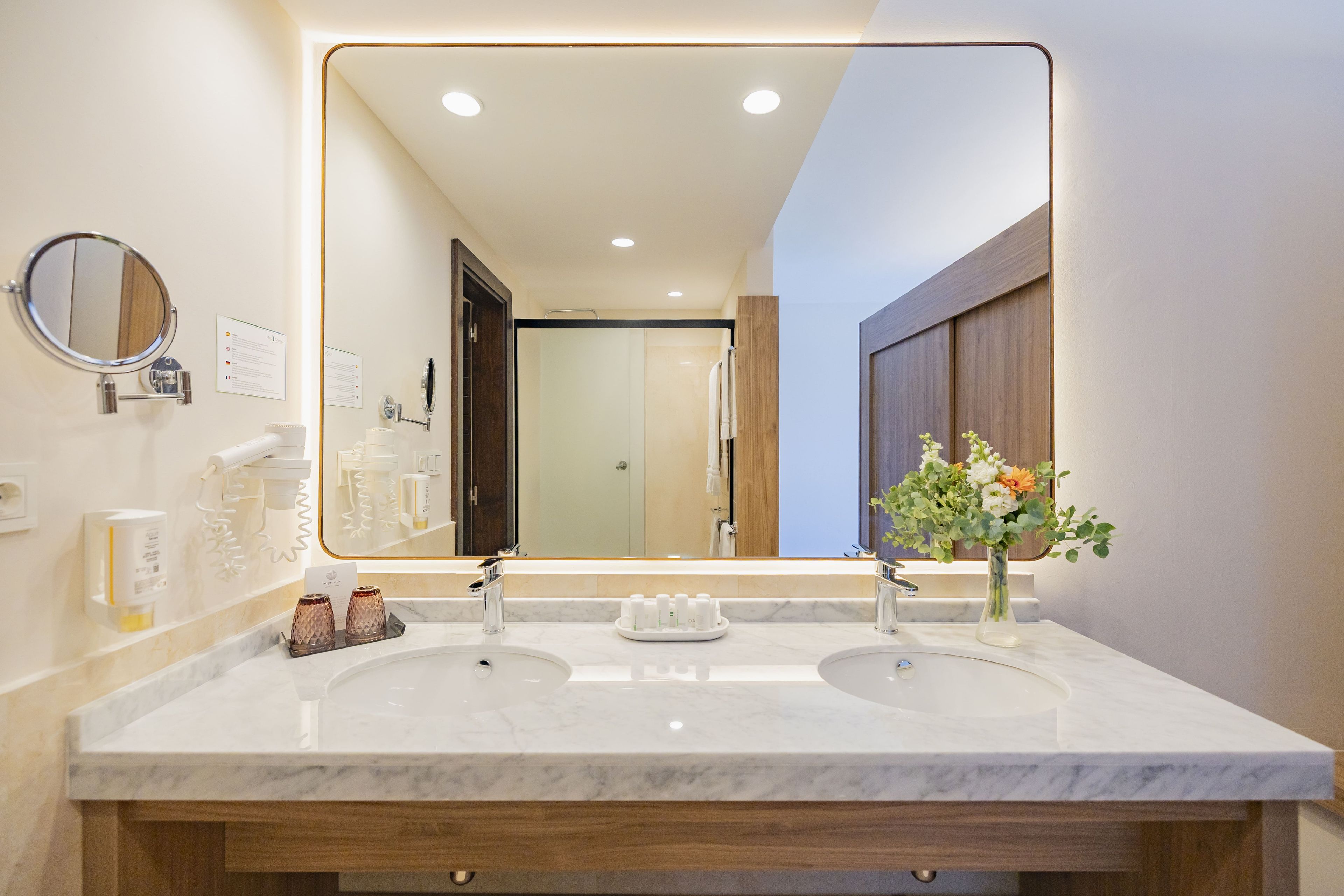 A modern bathroom with a double sink, a large mirror, and a vase of flowers.