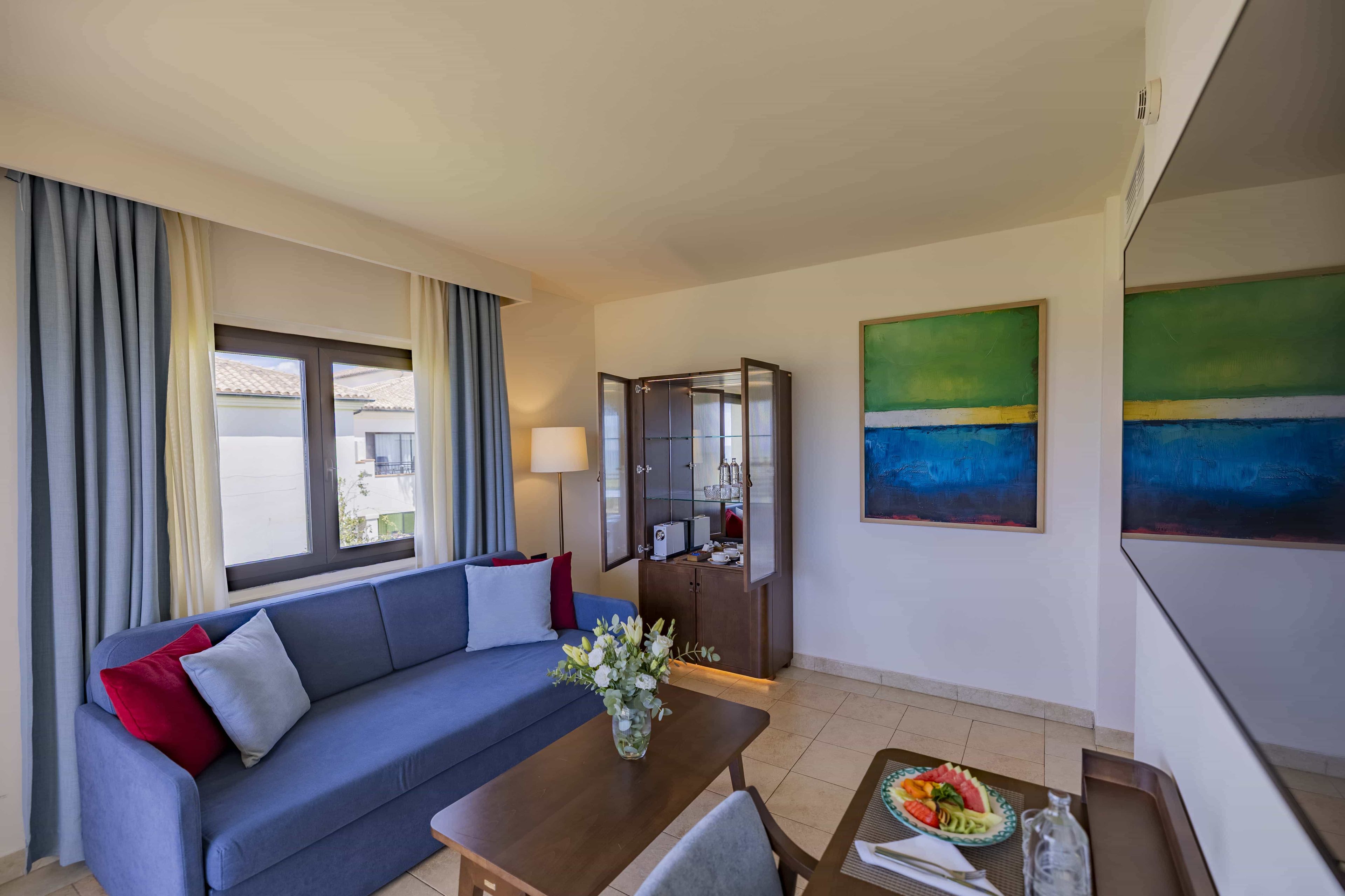 Een moderne hotelkamer met een blauwe bank, een eettafel met een bloemenvaas, een kast met een magnetron, en een groot schilderij aan de muur.