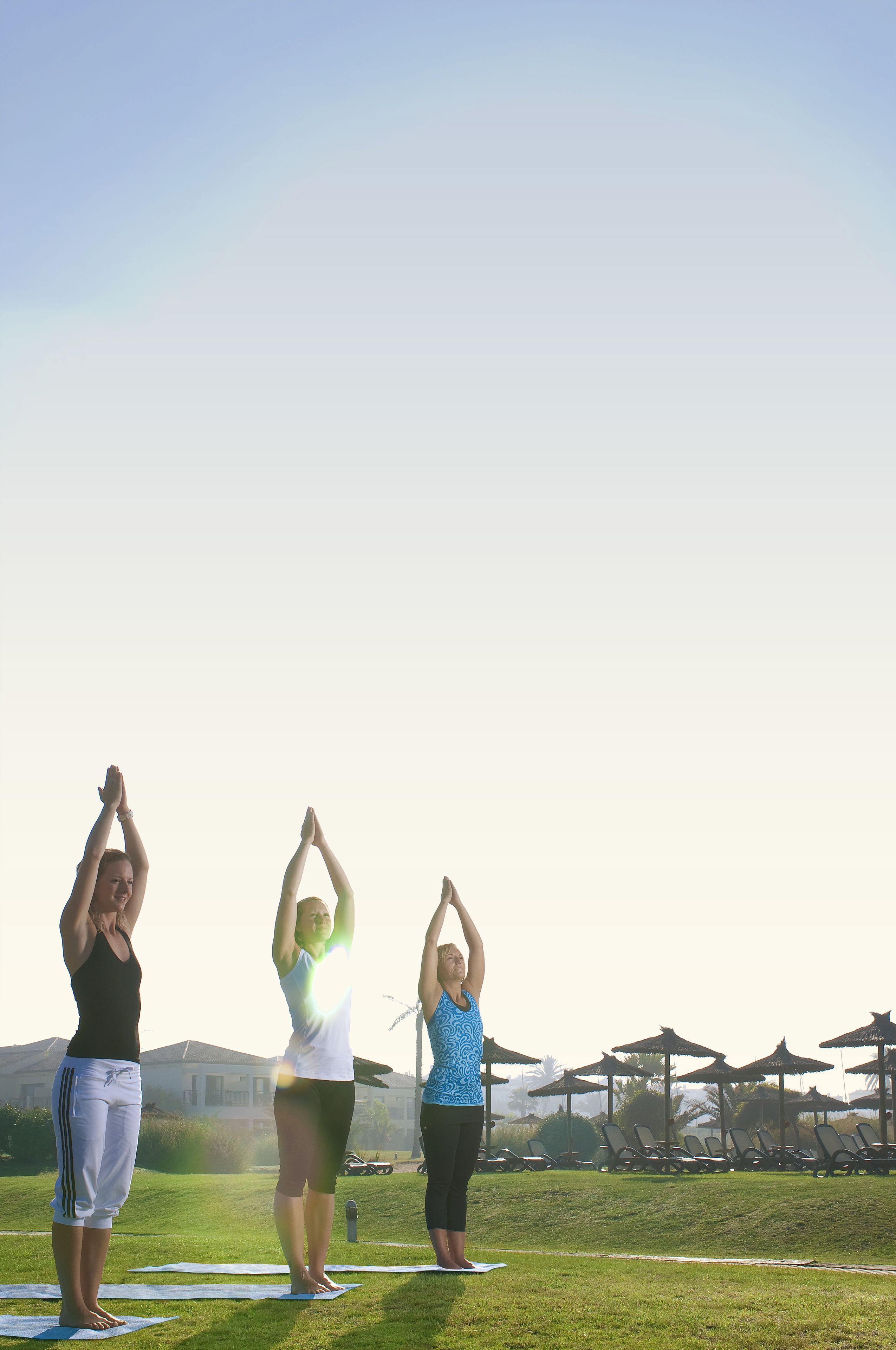 Three people doing yoga on mats in an outdoor setting with umbrellas and lounge chairs in the background.
