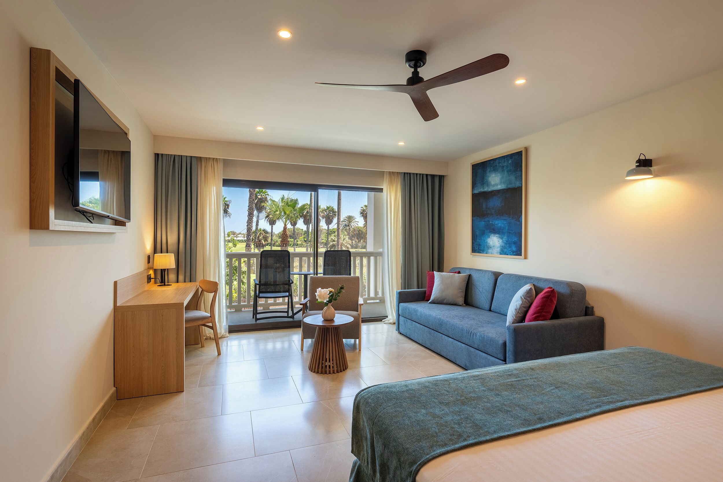Habitación de hotel con cama, escritorio, sillón, sofá, ventilador de techo y una ventana grande con cortinas.