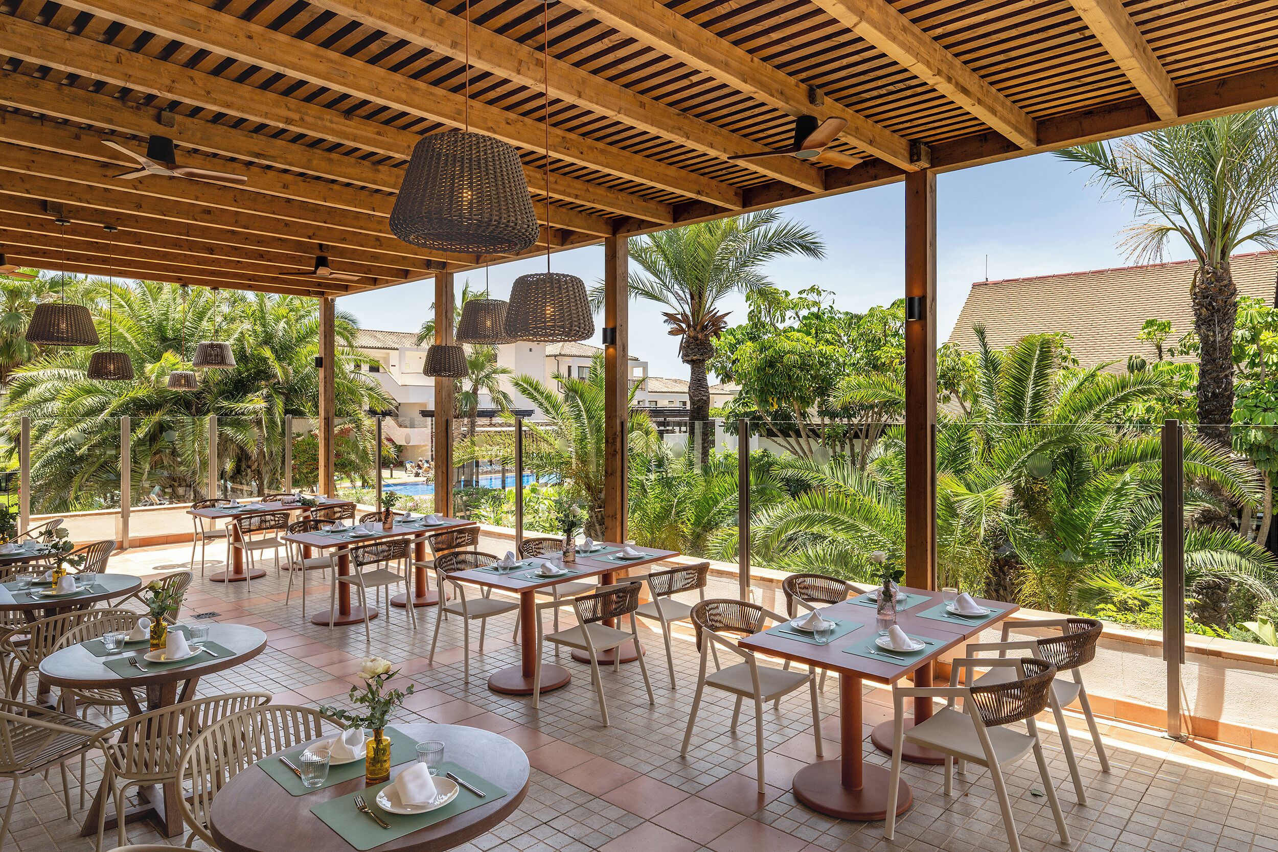 An outdoor dining area with tables and chairs, surrounded by greenery and a pool.