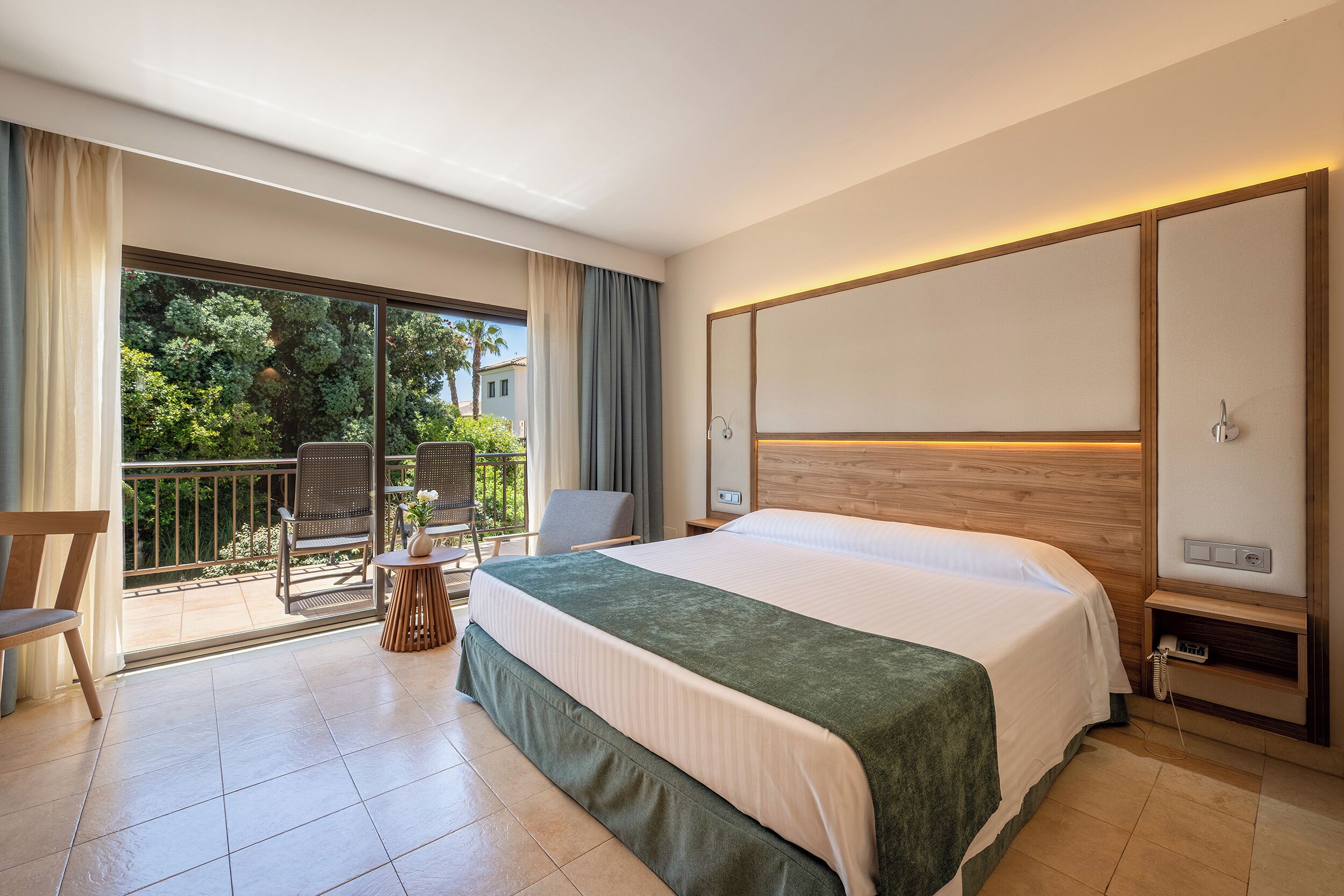 Habitación de hotel con cama, una mesita, una silla, un balcón con vistas a árboles y edificios, y cabecera de madera.