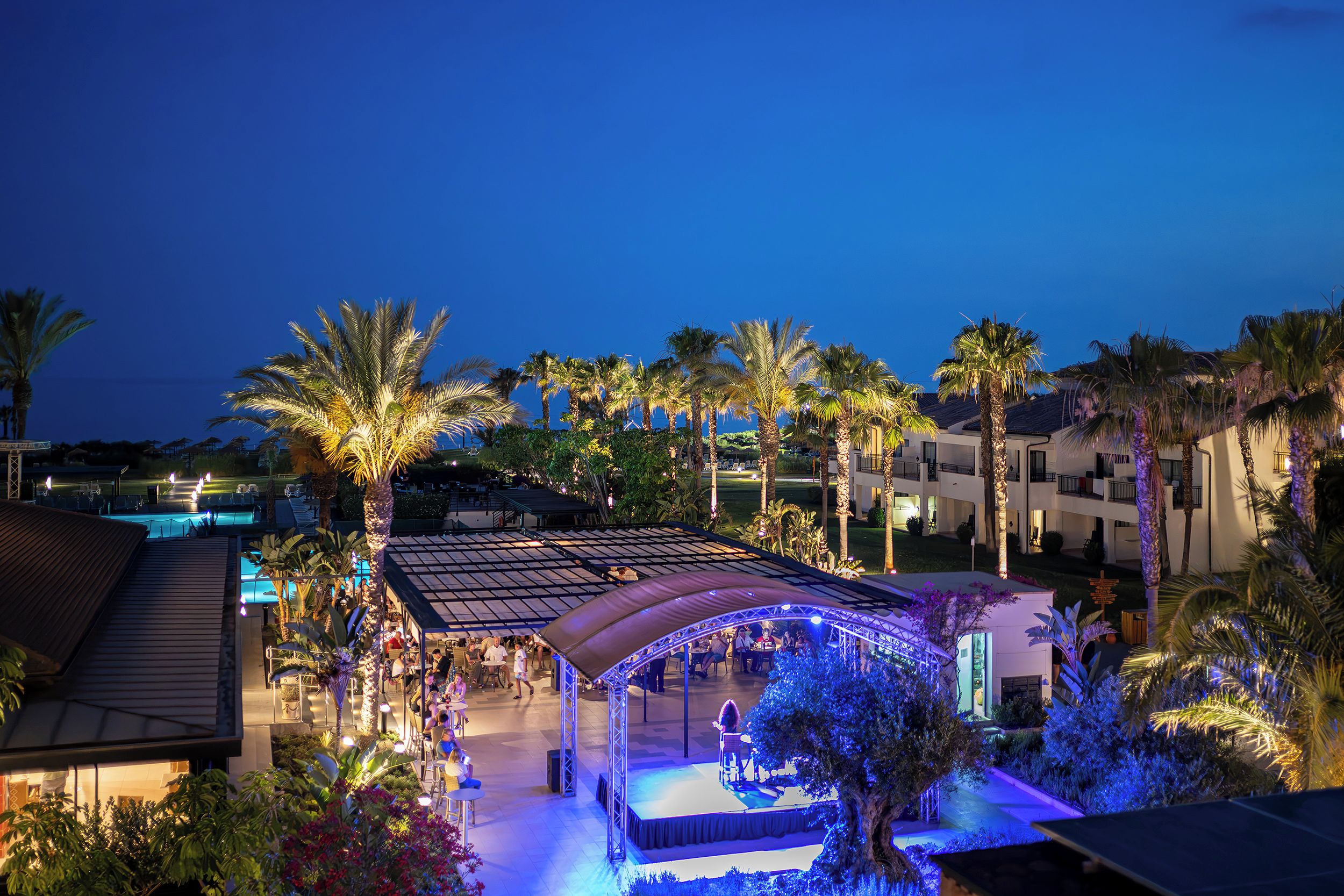 Un resort con piscina, palmeras y un espacio al aire libre cubierto donde hay gente.
