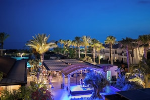 Un resort con piscina, palmeras y un espacio al aire libre cubierto donde hay gente.