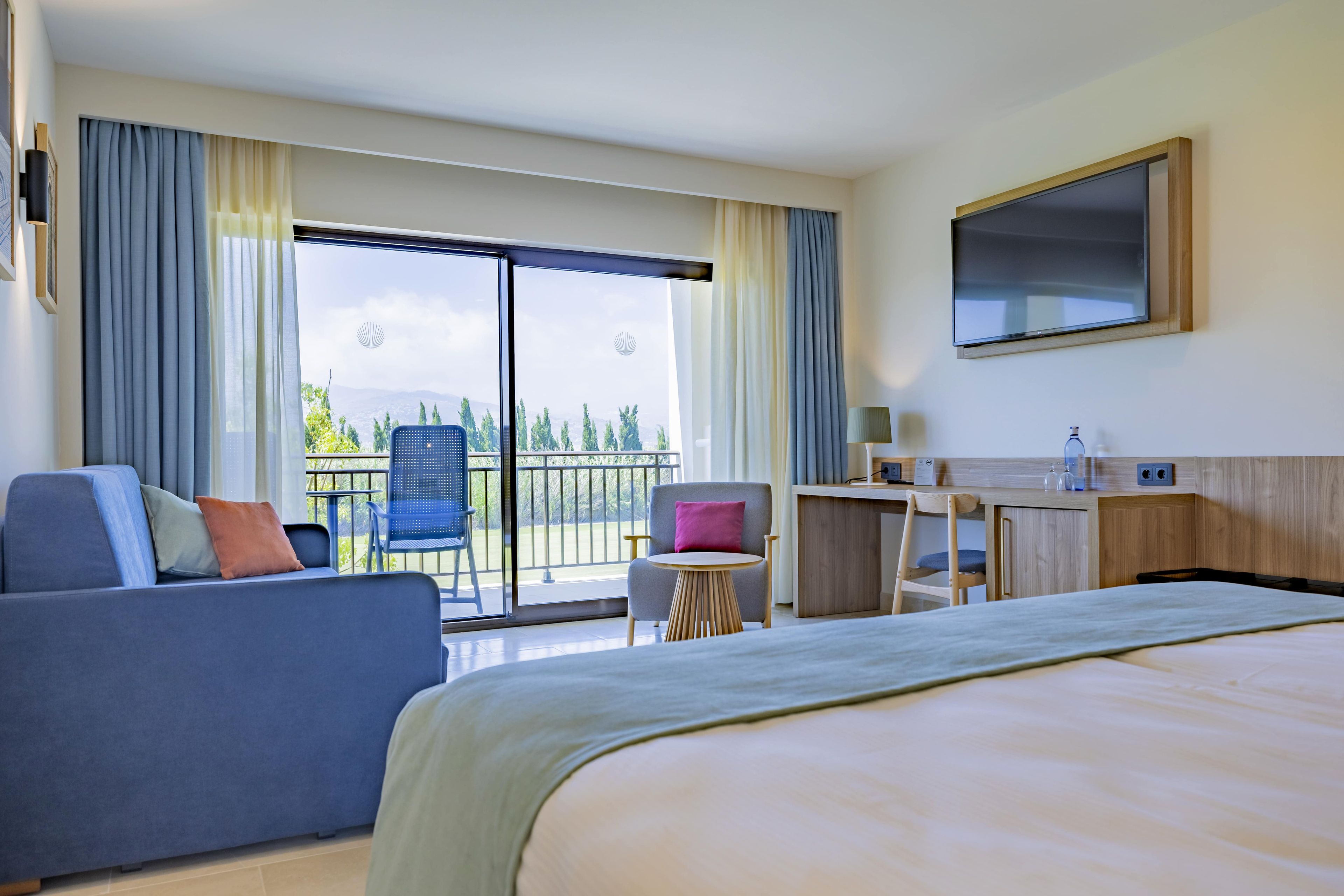 A hotel room with a bed, a sofa, a desk, a chair, and a balcony with a view.