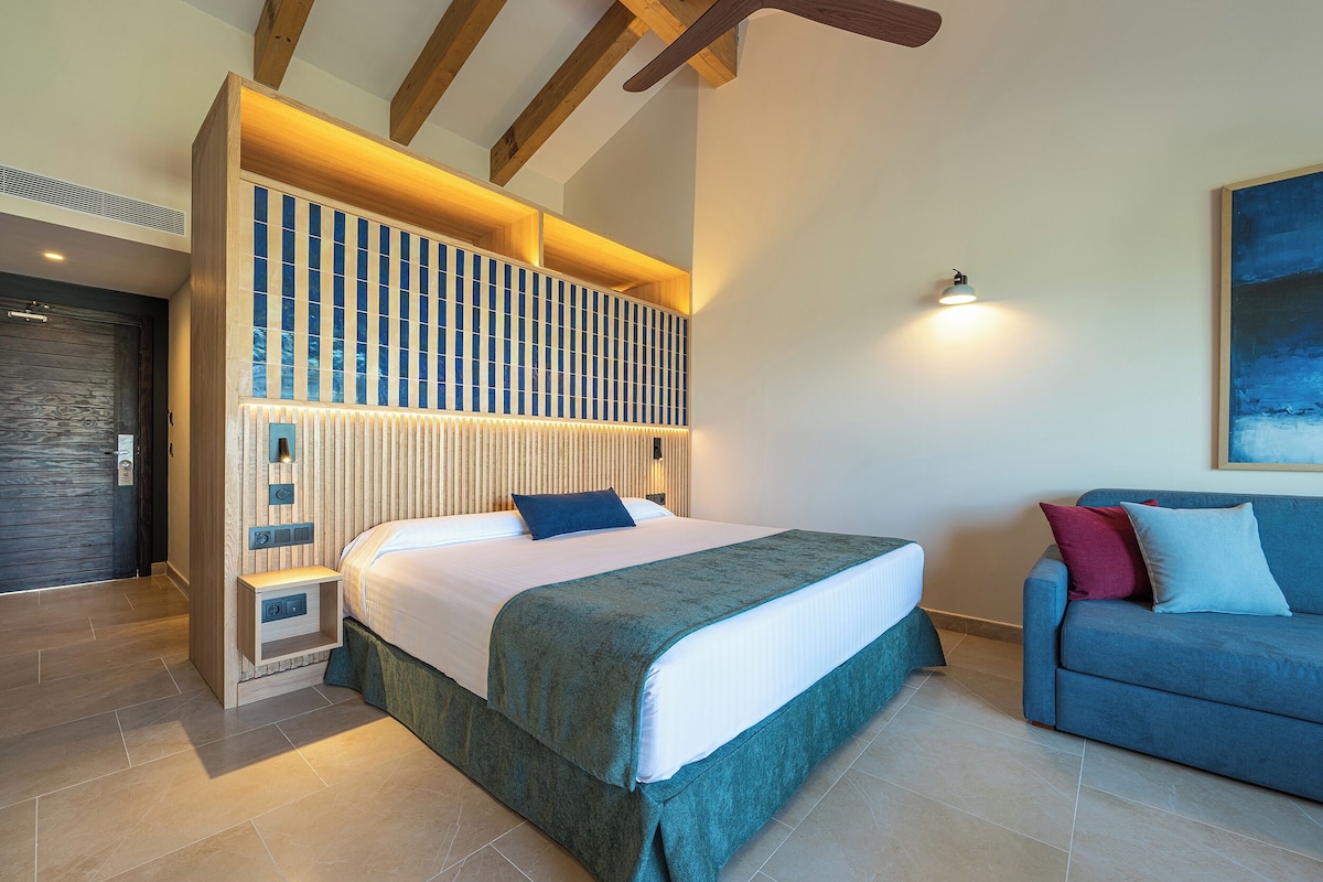 A modern hotel room with a large bed, a sofa, and a wall-mounted light fixture.