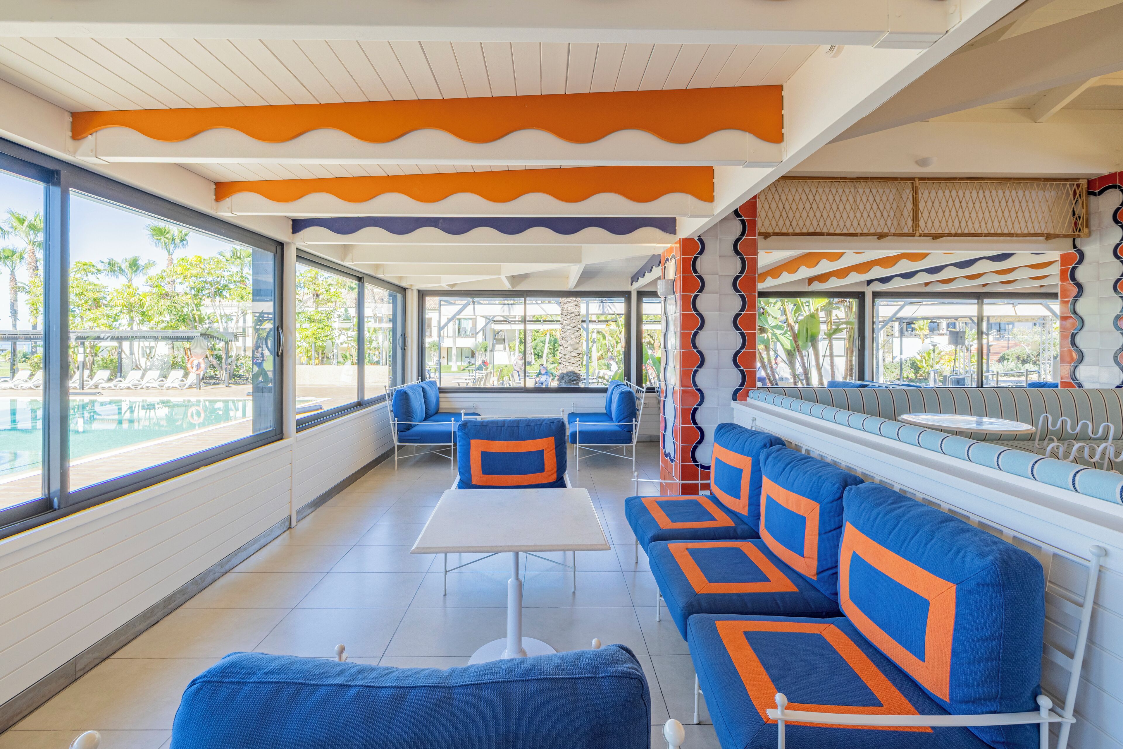 A modern lounge area with blue and orange seating, large windows, and a pool view.