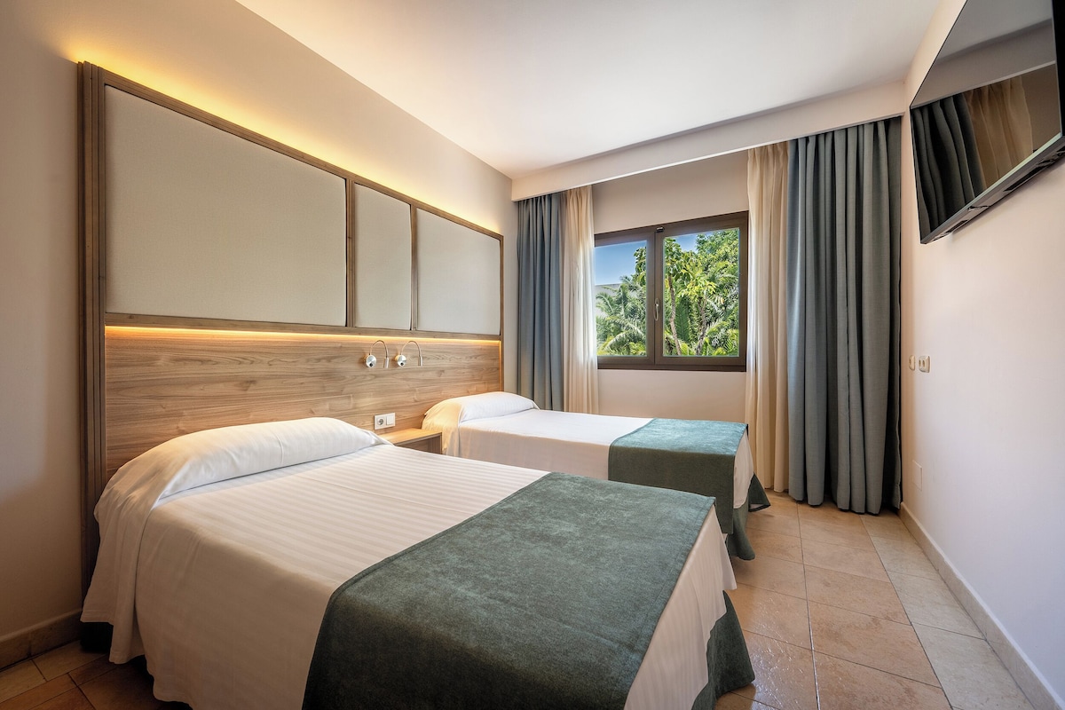A hotel room with two beds, a large window with curtains, and a flat-screen TV mounted on the wall.