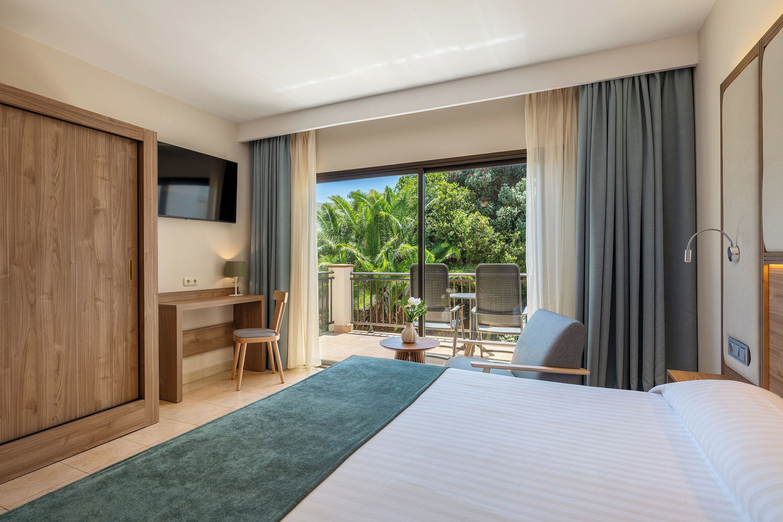 A hotel room with a bed, a desk, a chair, a TV, and a balcony with a view of greenery.