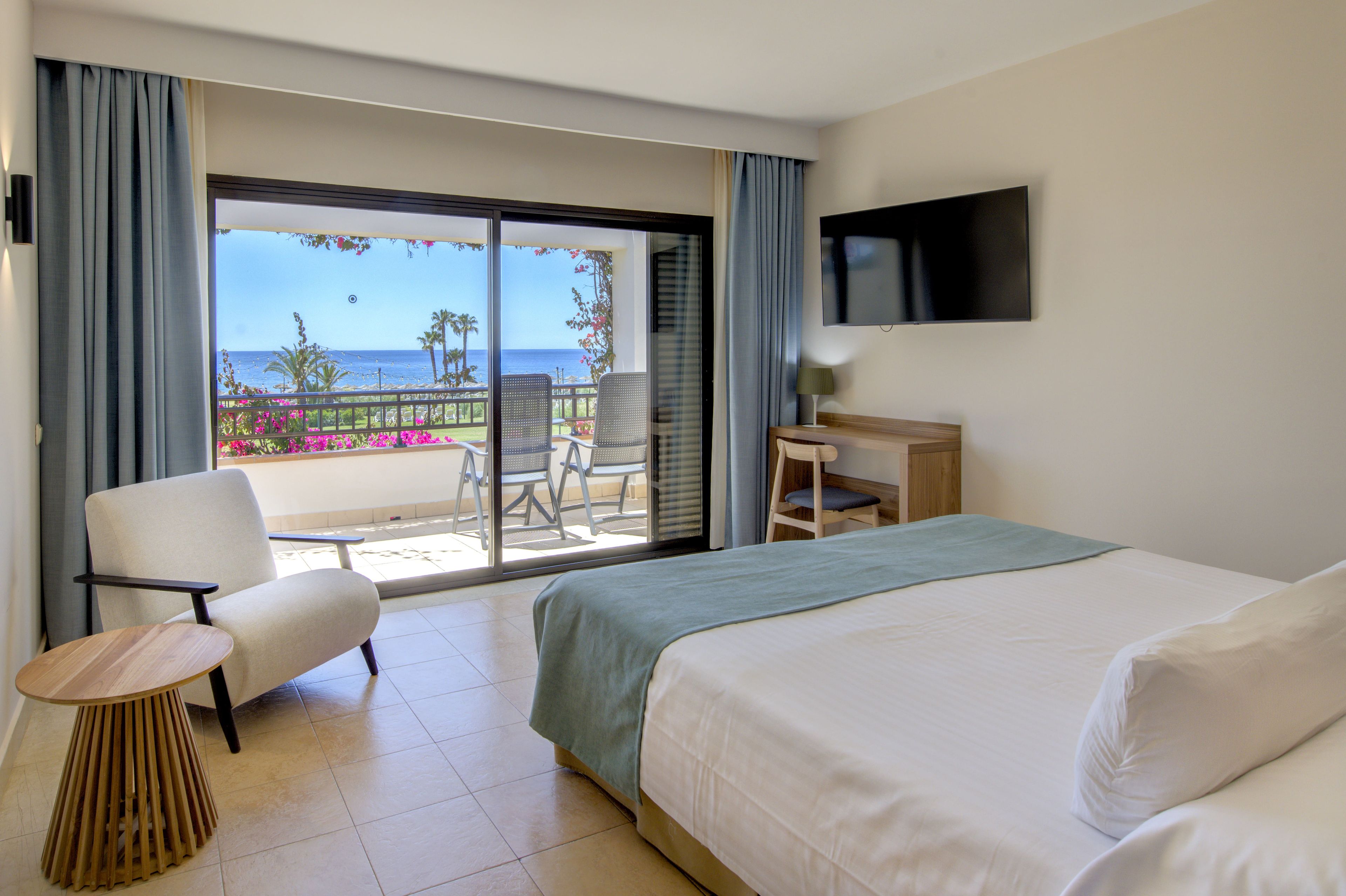 Een hotelkamer met een bed, een stoel, een tafeltje, een bureau, een televisie en een balkon met uitzicht op zee.