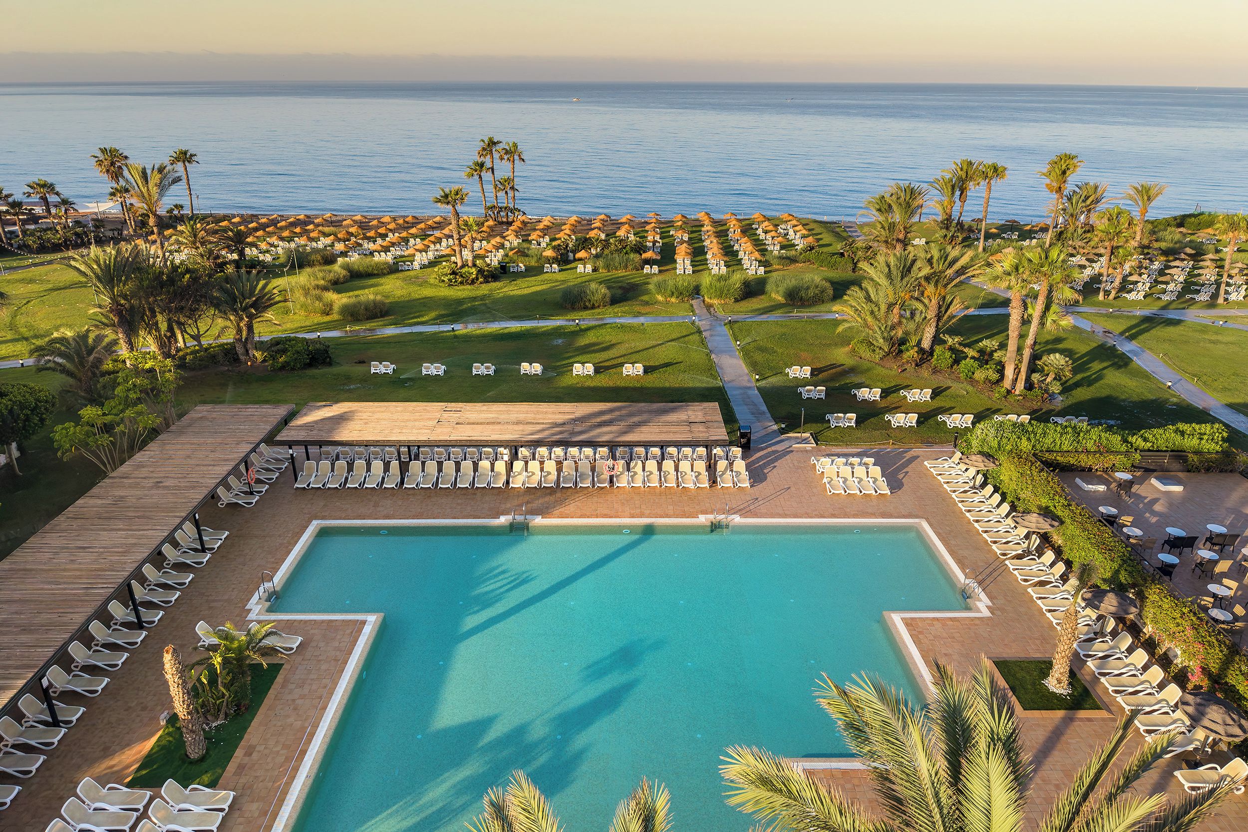 Un resort con una amplia piscina rodeada de sillas de descanso y palmeras, con vista a un mar tranquilo.