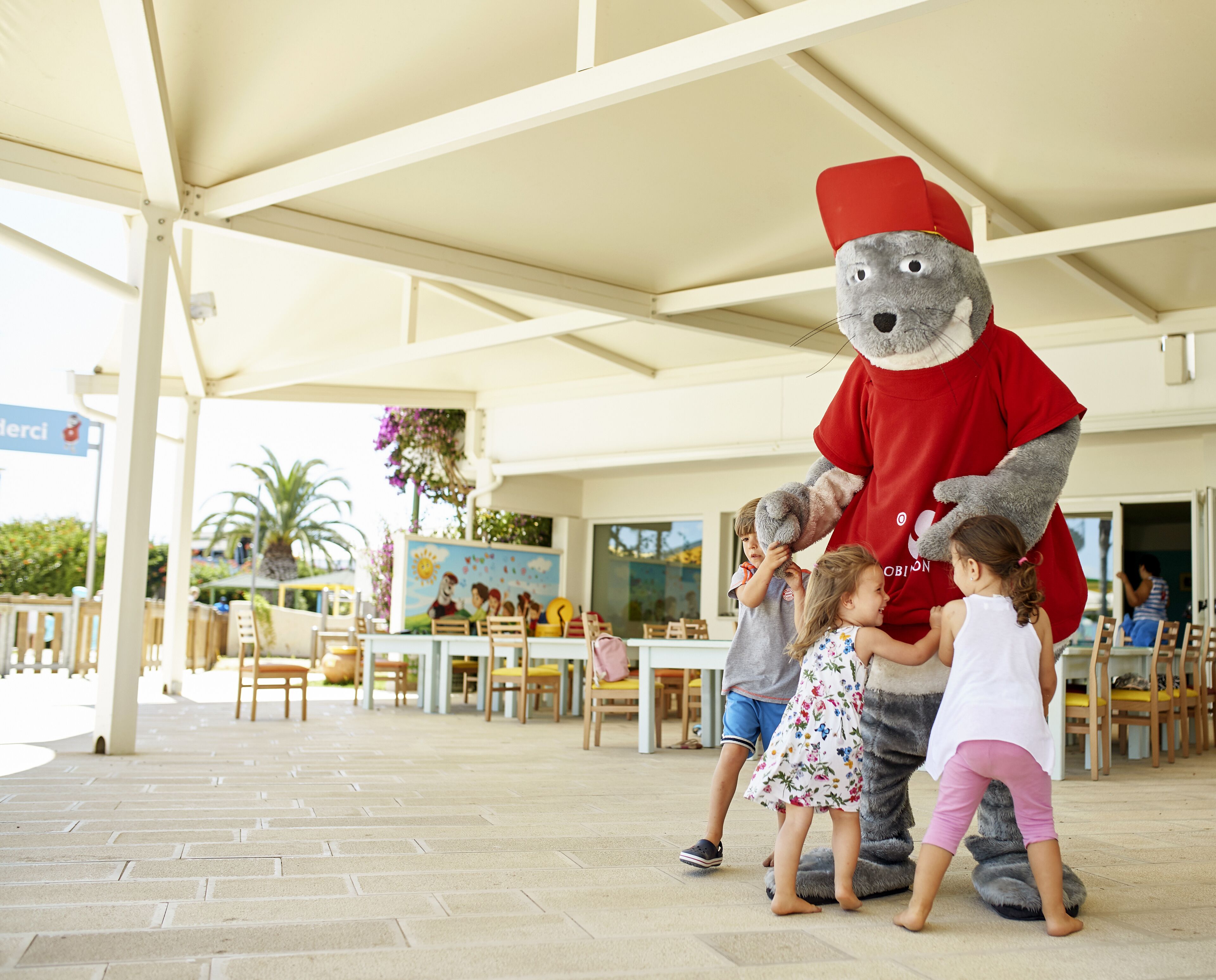 A mascot interacting with children in an outdoor seating area with tables and chairs, under a covered structure.