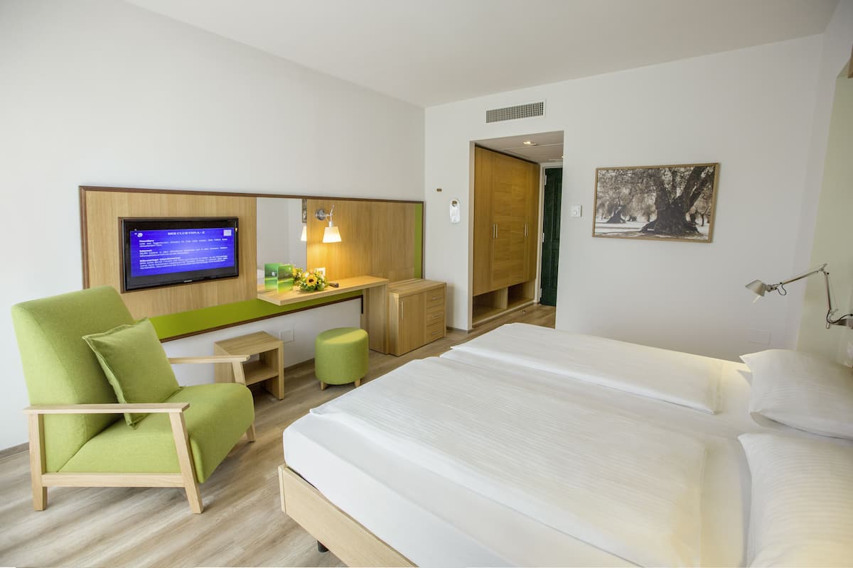 A hotel room with a bed, a desk with a computer, a green chair, and a small table.