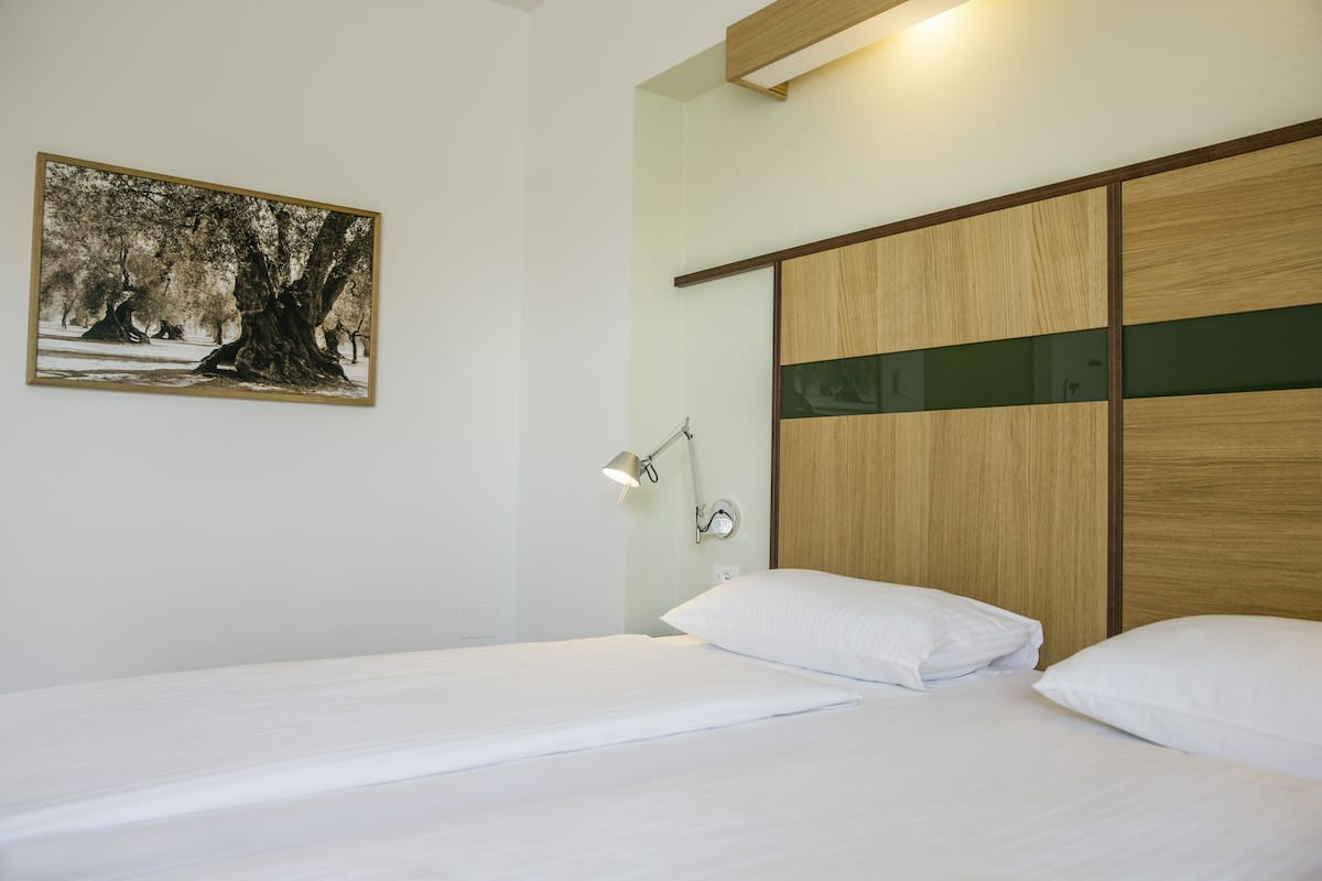 A bedroom with a bed, a nightstand, a wall-mounted lamp, and a framed picture of trees.
