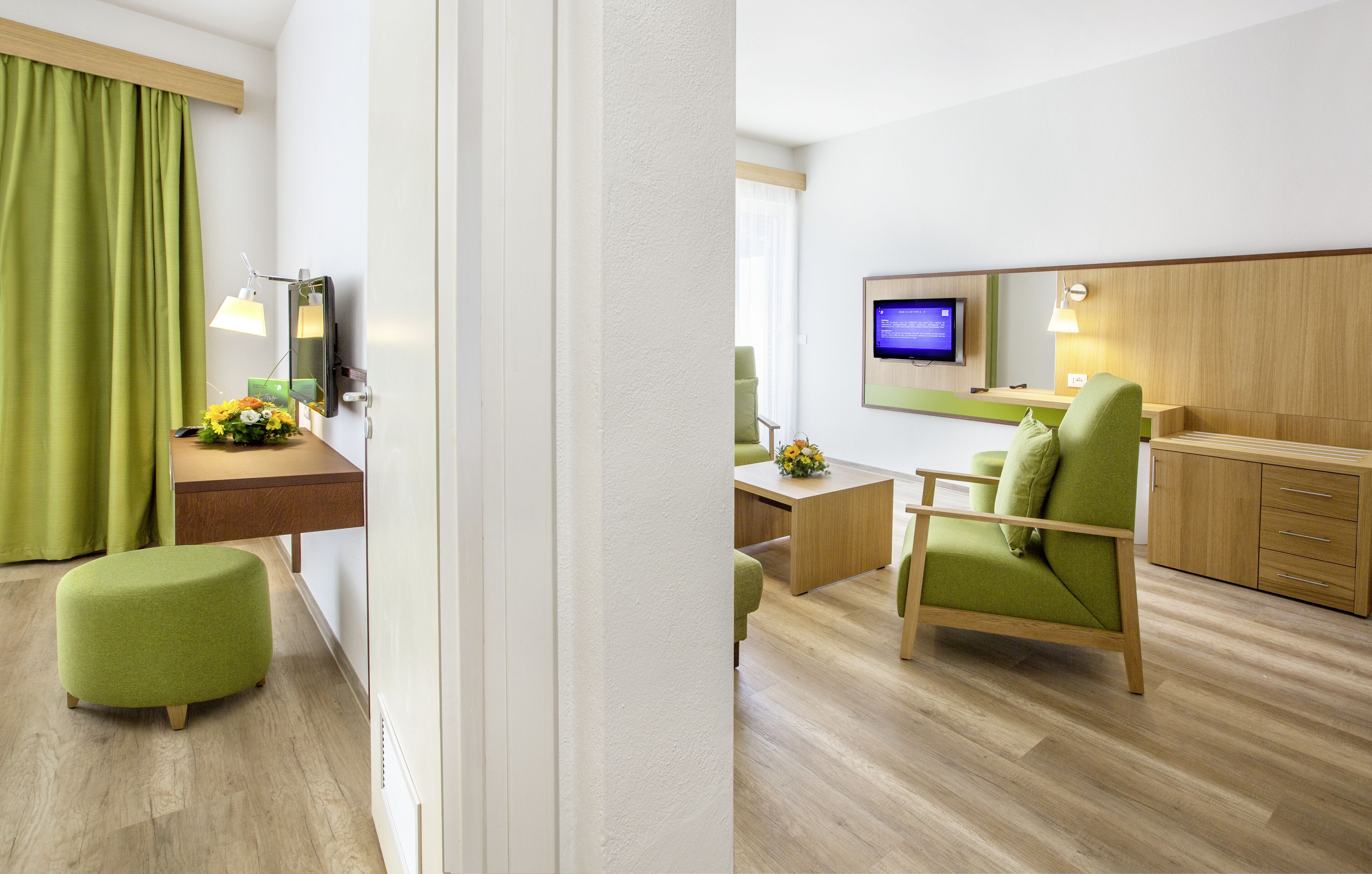 A hotel room with a wooden desk, a green armchair, a small ottoman, a TV, and a wooden cabinet.
