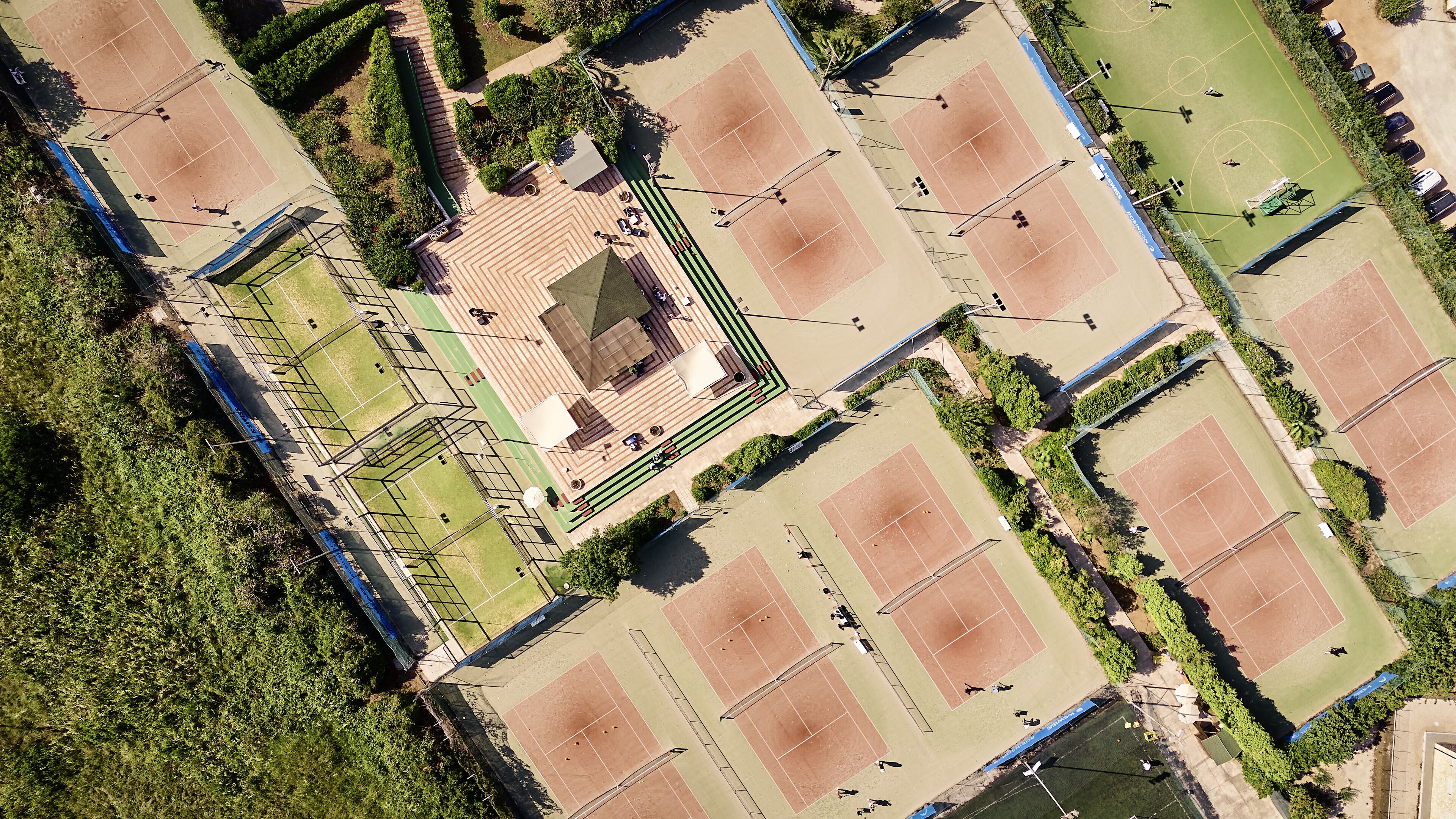 An aerial view of a sports complex with multiple tennis courts, a central pavilion, and surrounding greenery.