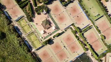 An aerial view of a sports complex with multiple tennis courts, a central pavilion, and surrounding greenery.