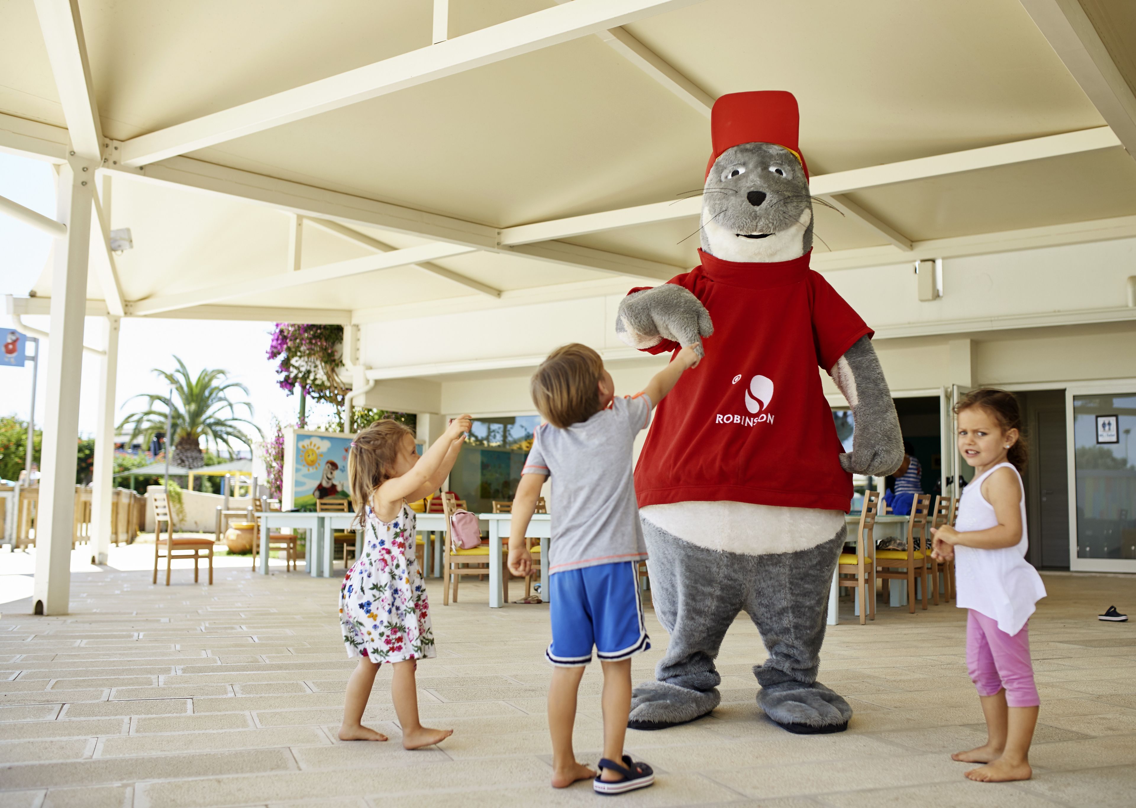 Une mascotte, vêtue d’un t-shirt et d’un chapeau rouges, interagit avec des enfants dans un espace extérieur aménagé avec des tables et des chaises.