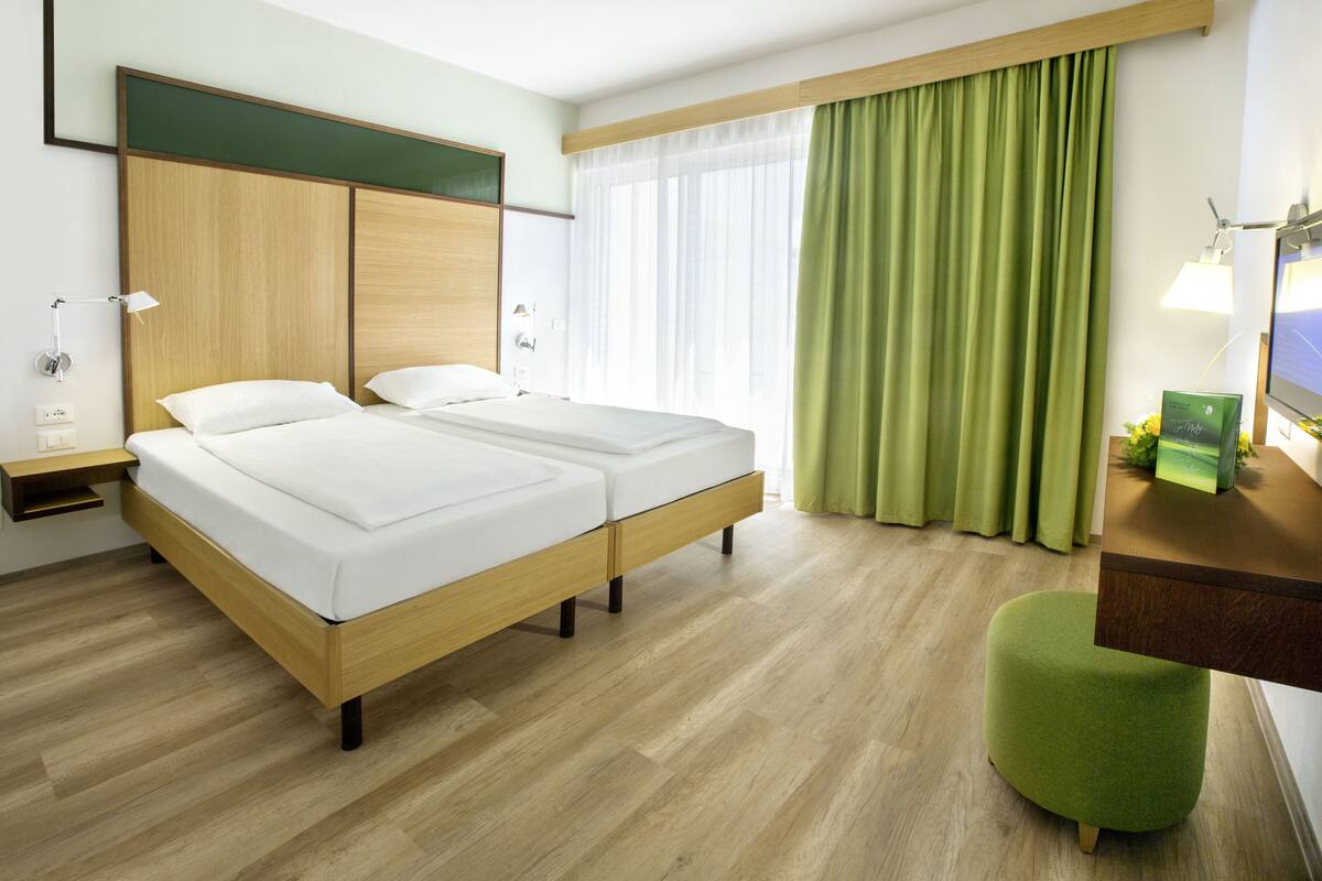 A hotel room with a double bed, a desk with a computer, a green ottoman, and a large window with green curtains.