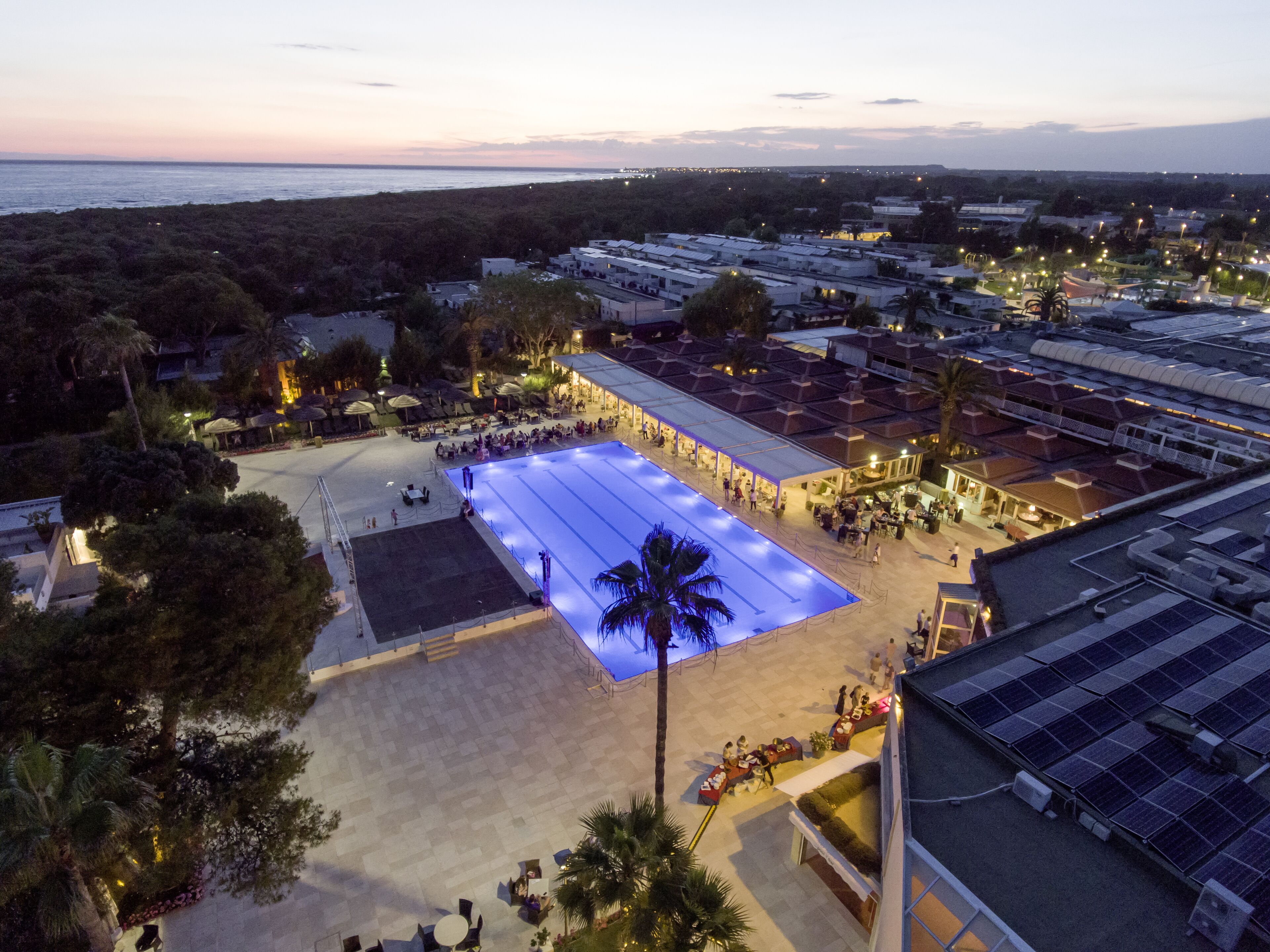 Eine Luftaufnahme eines Resorts mit einem großen Schwimmbecken, mehreren Speisebereichen und Blick auf das Meer im Abenddämmerung.