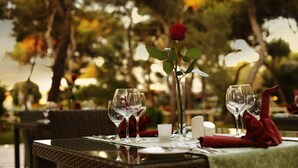 Una mesa de comedor al aire libre con una rosa en un florero, copas de vino y un mantel individual rojo.