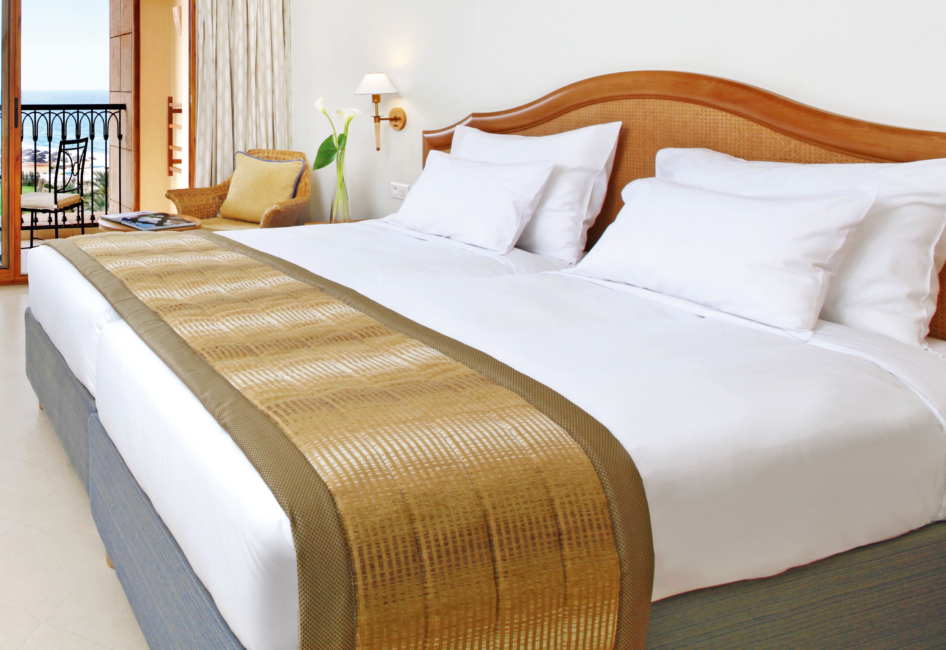 A large bed with white linens and a beige accent runner, a wooden headboard, and a nightstand with a lamp and a vase of flowers.