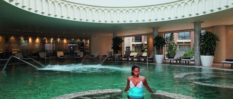 Una amplia piscina interior con un diseño de mosaico central, rodeada de sillones de descanso y plantas en macetas, bajo un techo de cristal que ofrece vistas del exterior del edificio.