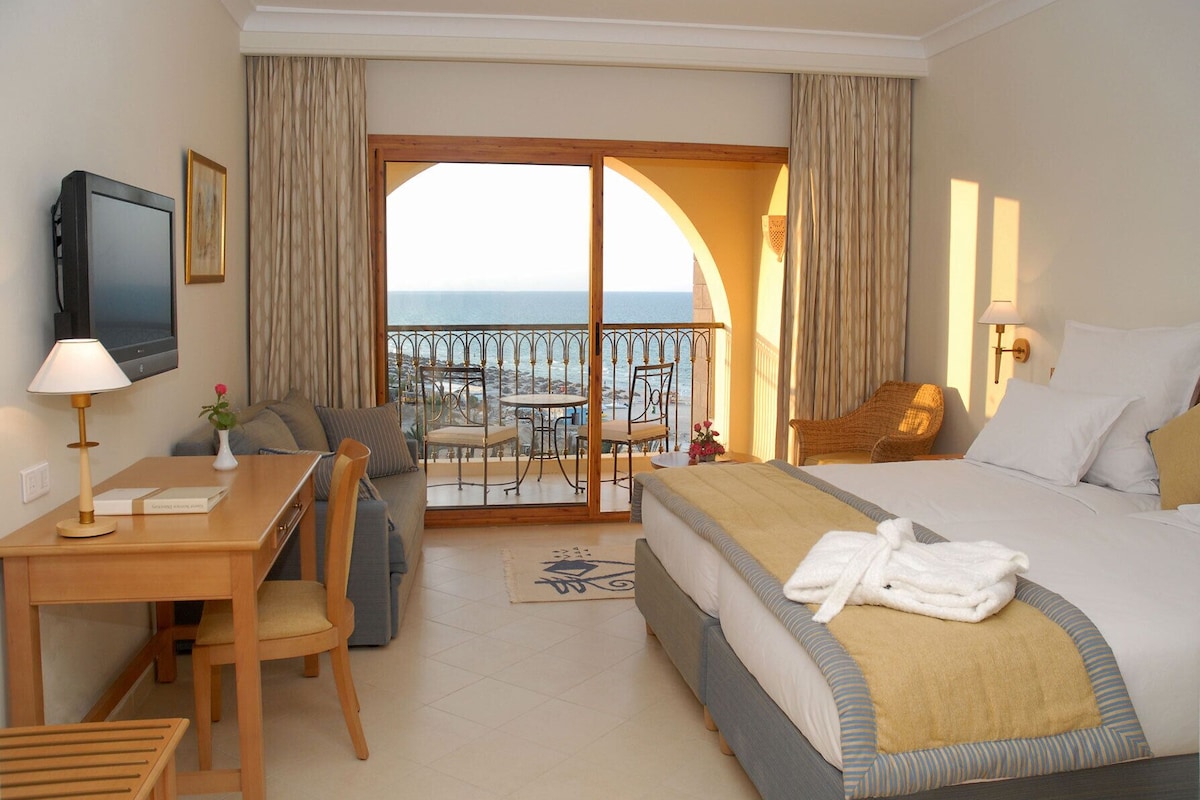 A hotel room with a bed, a desk, a chair, a TV, and a balcony with a view of the sea.