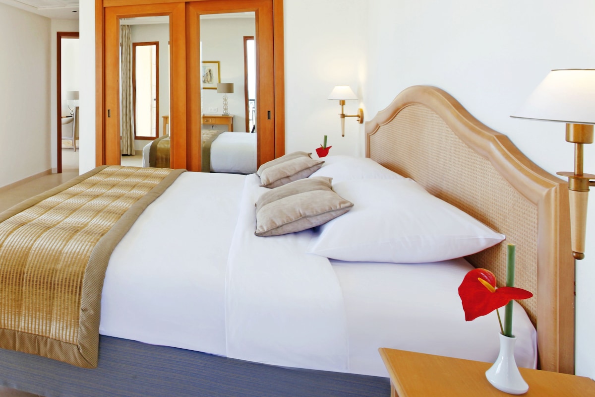 A hotel room with a large bed, a bedside table with a lamp, and a view of the hallway.