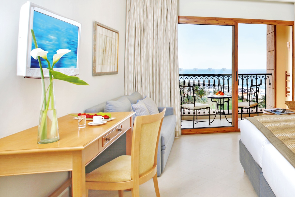 A hotel room with a balcony, a dining table with chairs, a sofa, and a bed.