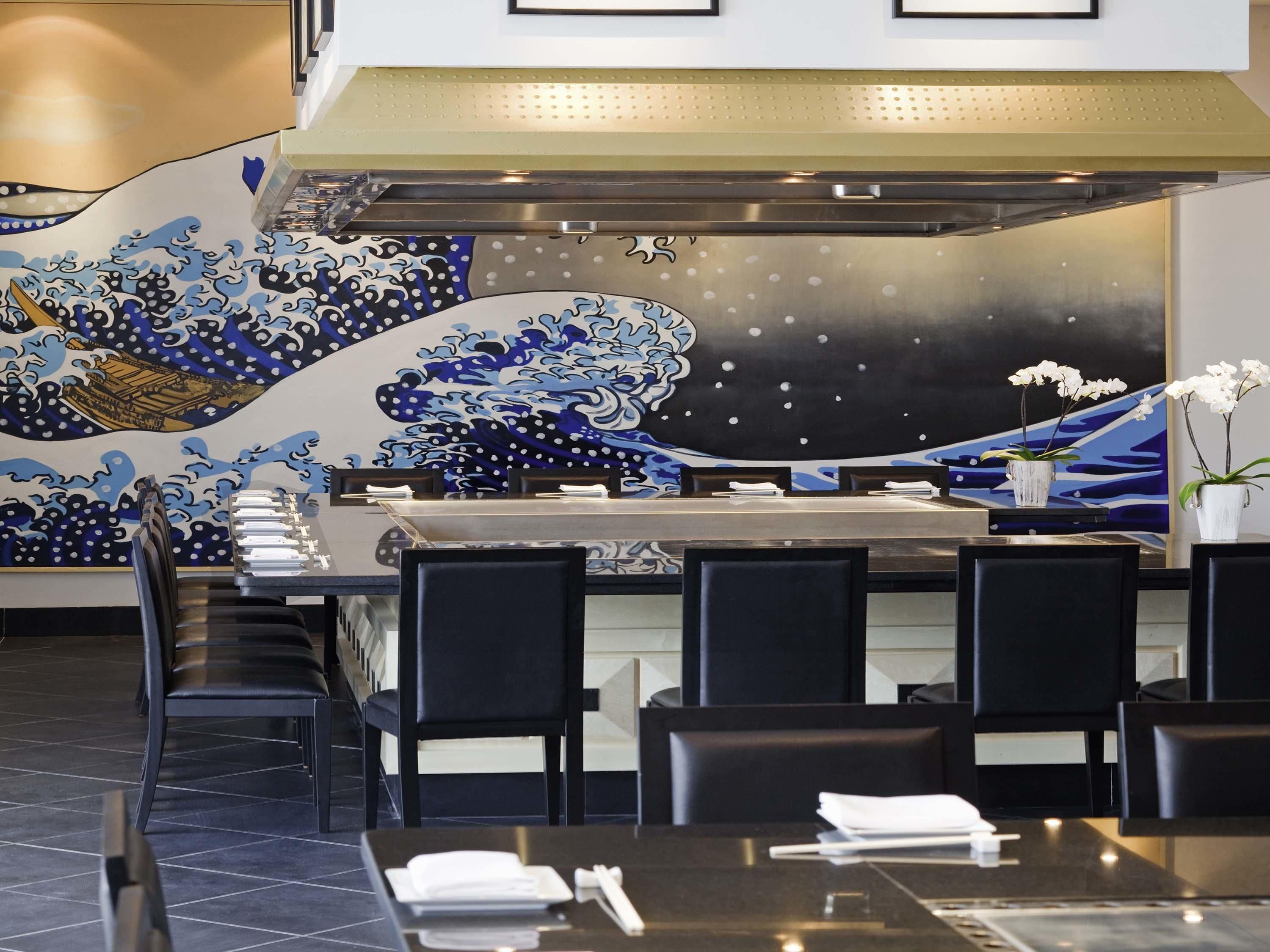 Um restaurante com um balcão de sushi, cadeiras pretas e uma parede decorativa com padrão de ondas.