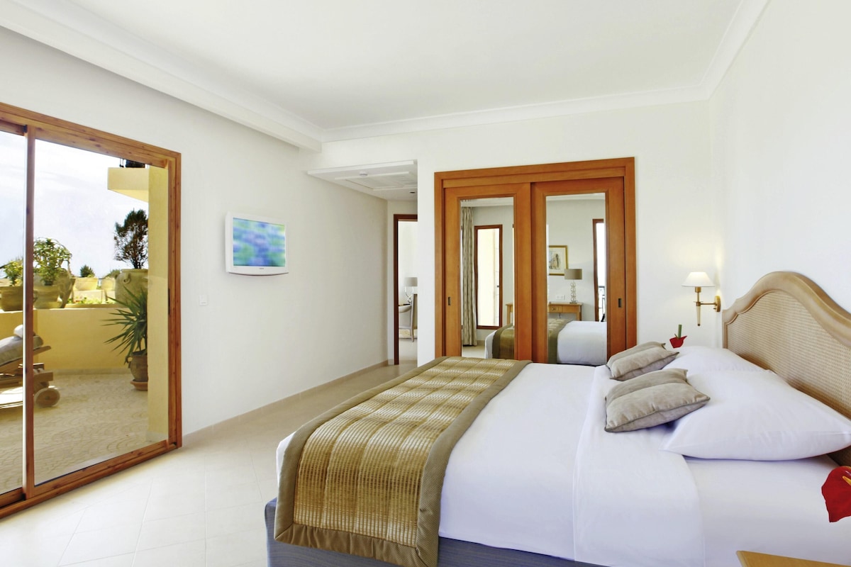 A hotel room with a bed, a bedside table, a lamp, a painting, and a view through a glass door.