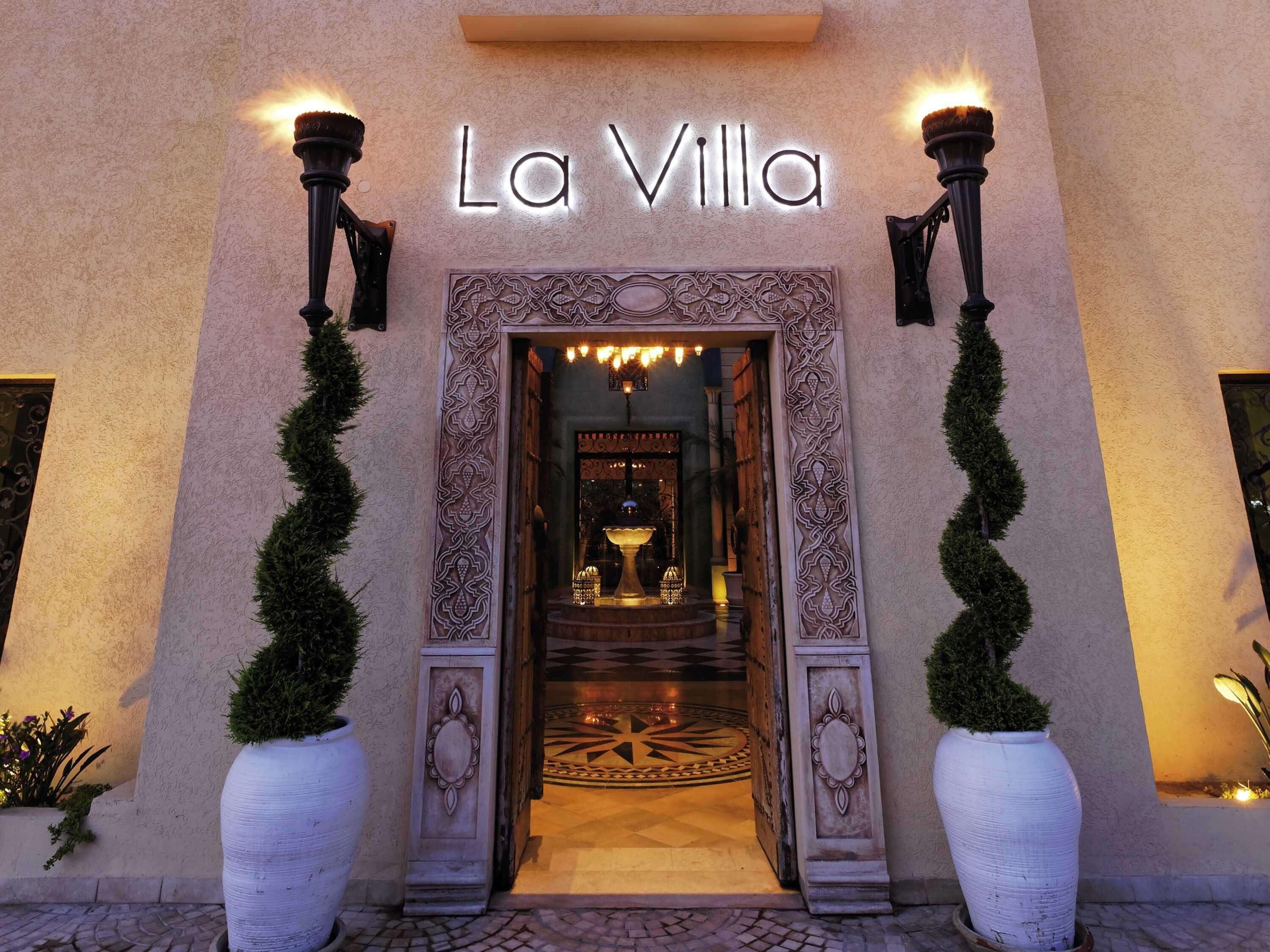 The entrance of La Villa hotel with a decorative archway, a sign above the entrance, and two large potted plants flanking the doorway.