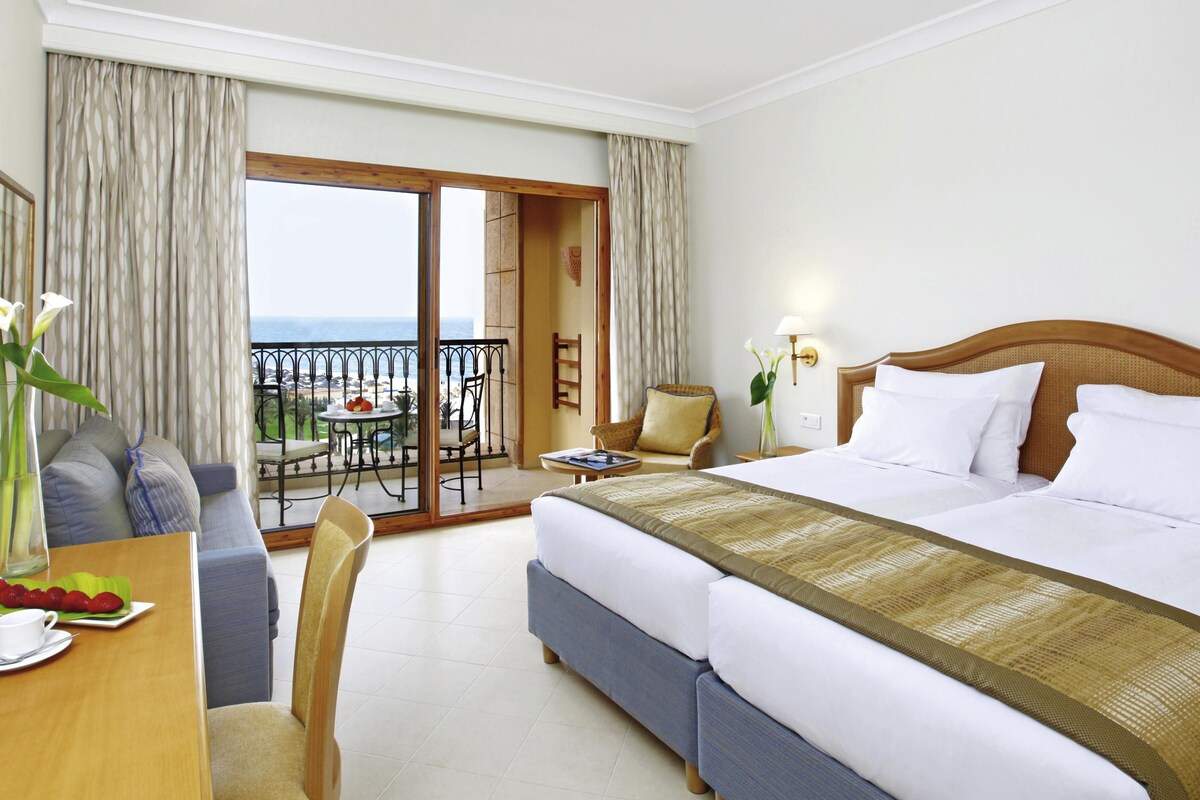 A hotel room with a large bed, a balcony with a table and chairs, and a view of the sea.