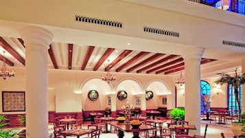 Un amplio vestíbulo de hotel con varias zonas de restauración, techos altos con vigas de madera y un balcón con iluminación azul.
