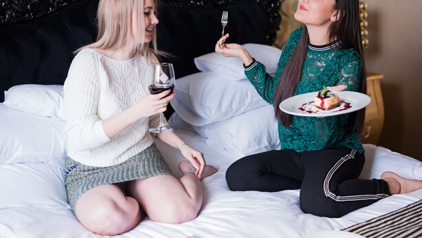 Two people sitting on a bed with a black headboard, one holding a wine glass and the other holding a plate with food. - Weekend Boutique Hotel (Chisinau)