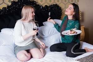 Dos personas sentadas en una cama con cabecera negra, una sosteniendo una copa de vino y la otra, un plato con comida.
