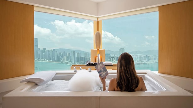 Une personne se détend dans une baignoire, avec vue sur la ville, une serviette et un livre à portée de main.