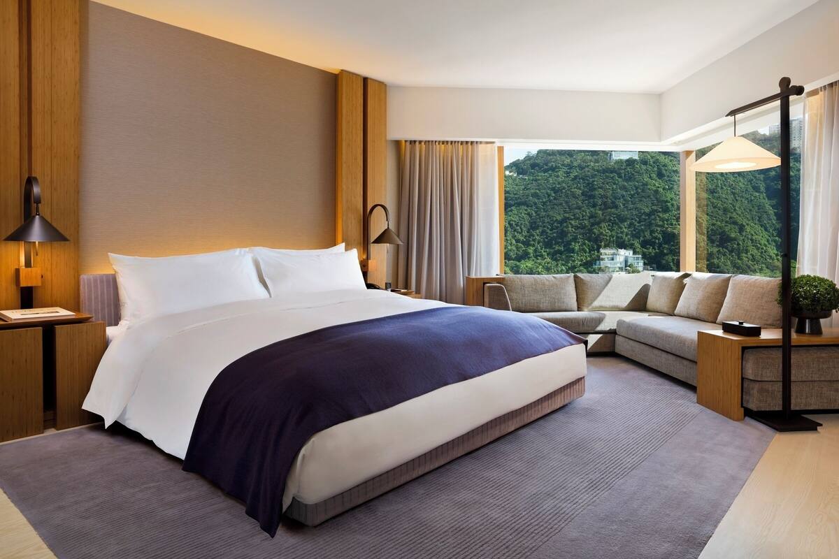 A modern hotel room with a large bed, a sofa, a desk, and a view of a forested area.