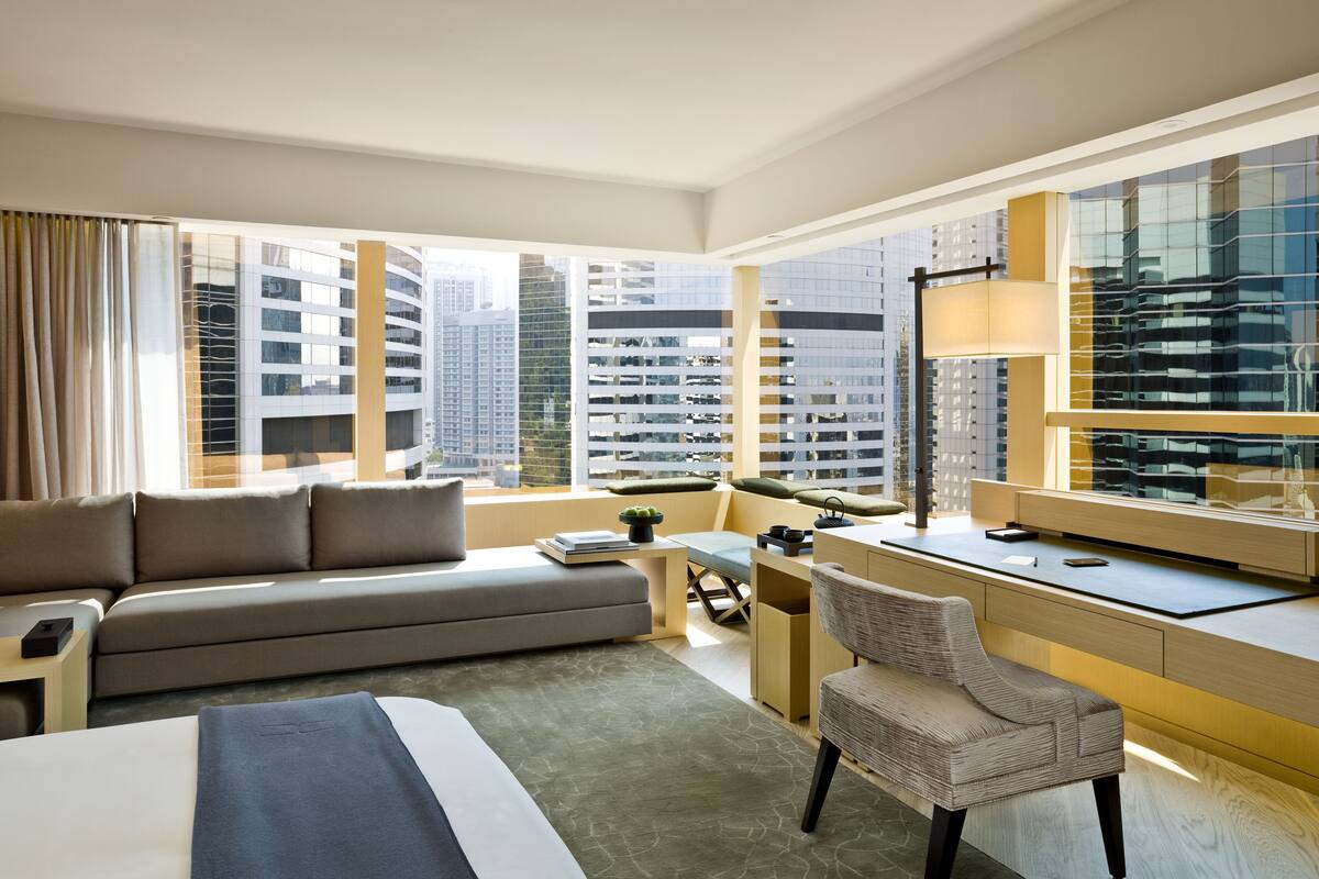 A modern hotel room with a large bed, a sofa, a desk, and a chair. There is a large window with a view of tall buildings.