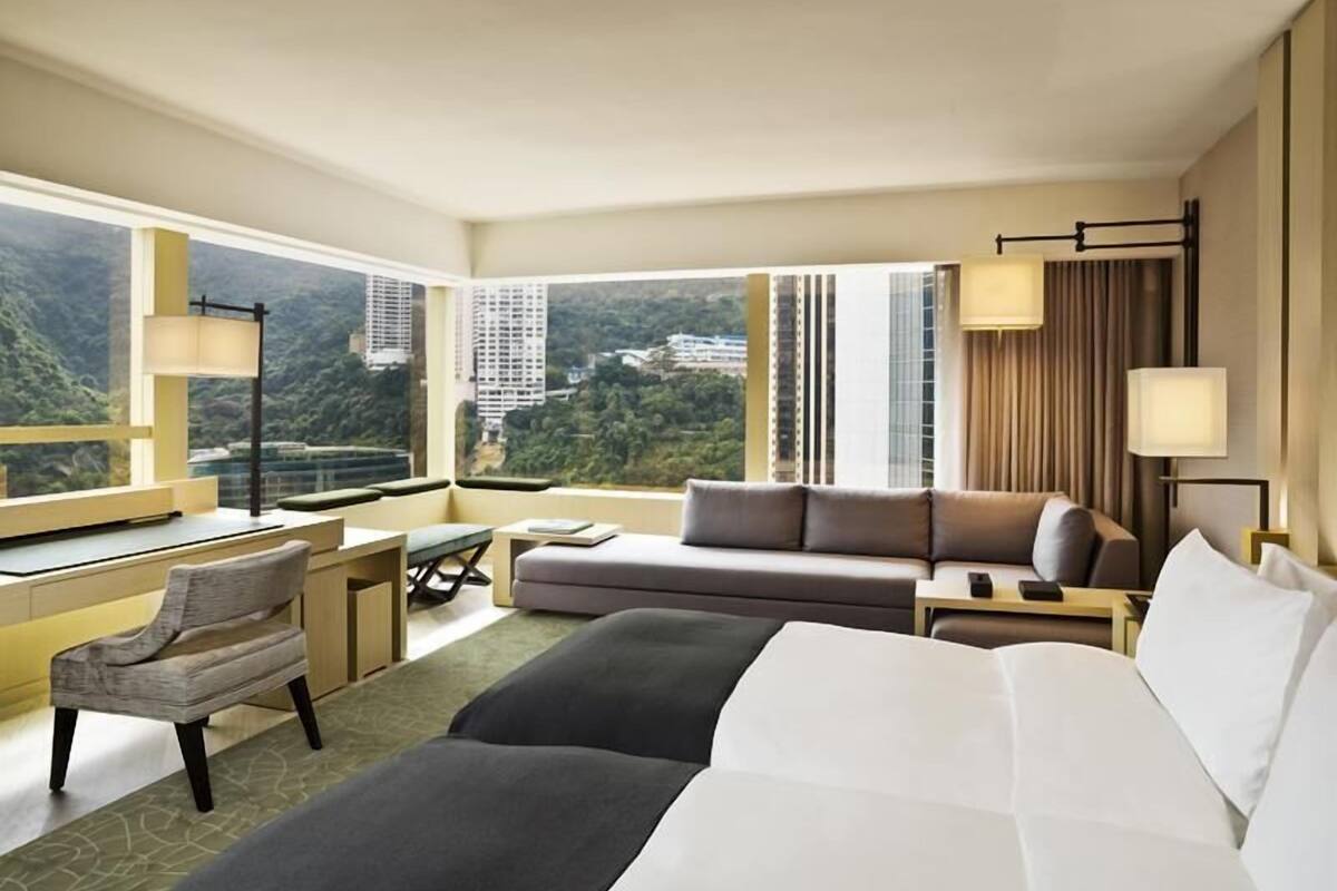 A hotel room with a large bed, a sofa, a desk, and a chair. There are large windows with a view of buildings and greenery.