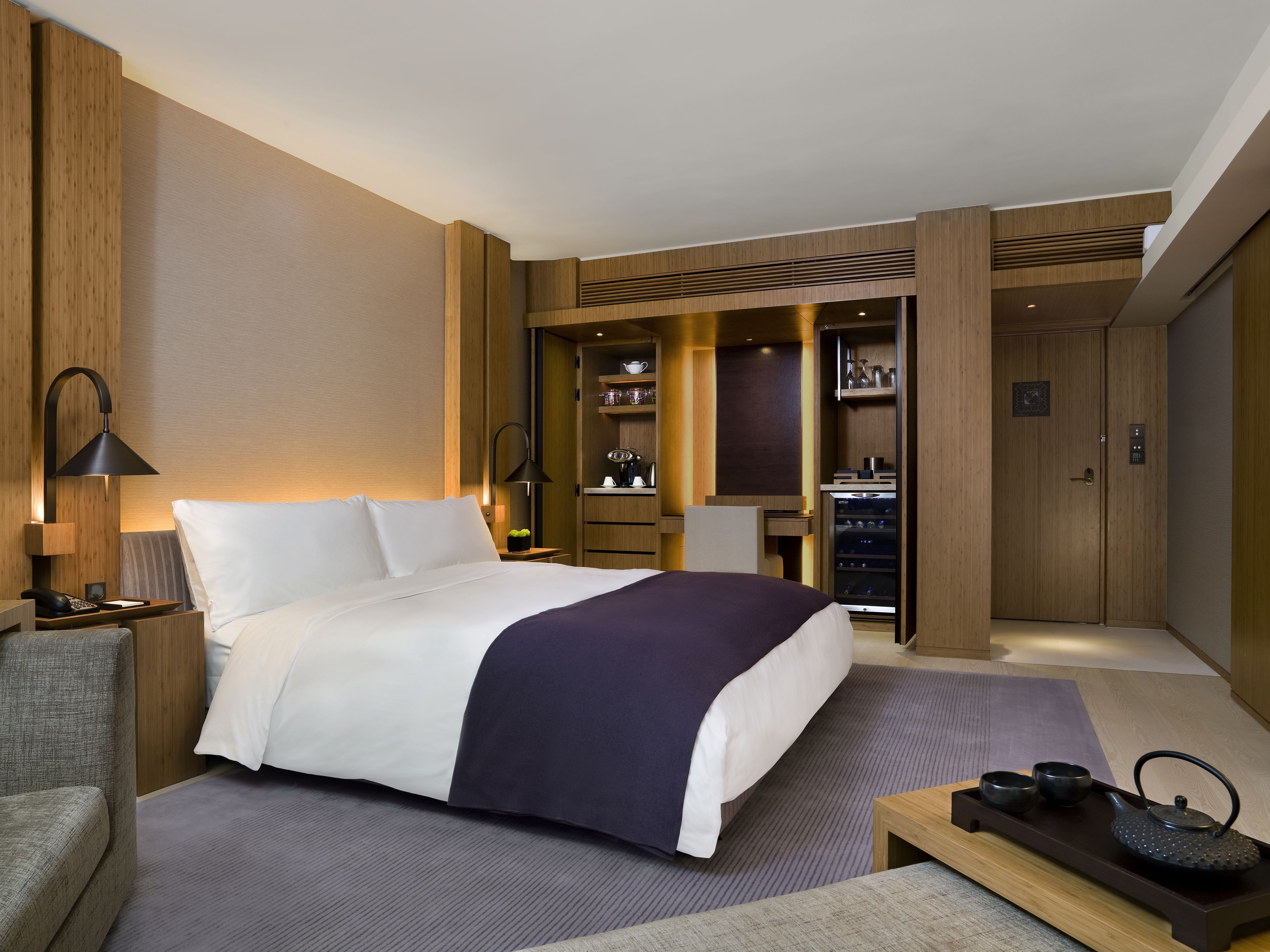 A modern hotel room with a large bed, a desk, a chair, a wardrobe, and a minibar.