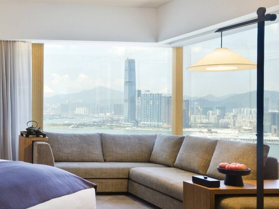 A modern hotel room with a large window offering a city view, a sofa, a bedside table, and a lamp.