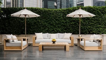 A modern outdoor seating area with a sofa, two armchairs, and a coffee table, all with wooden frames and light-colored cushions. There are two umbrellas providing shade.