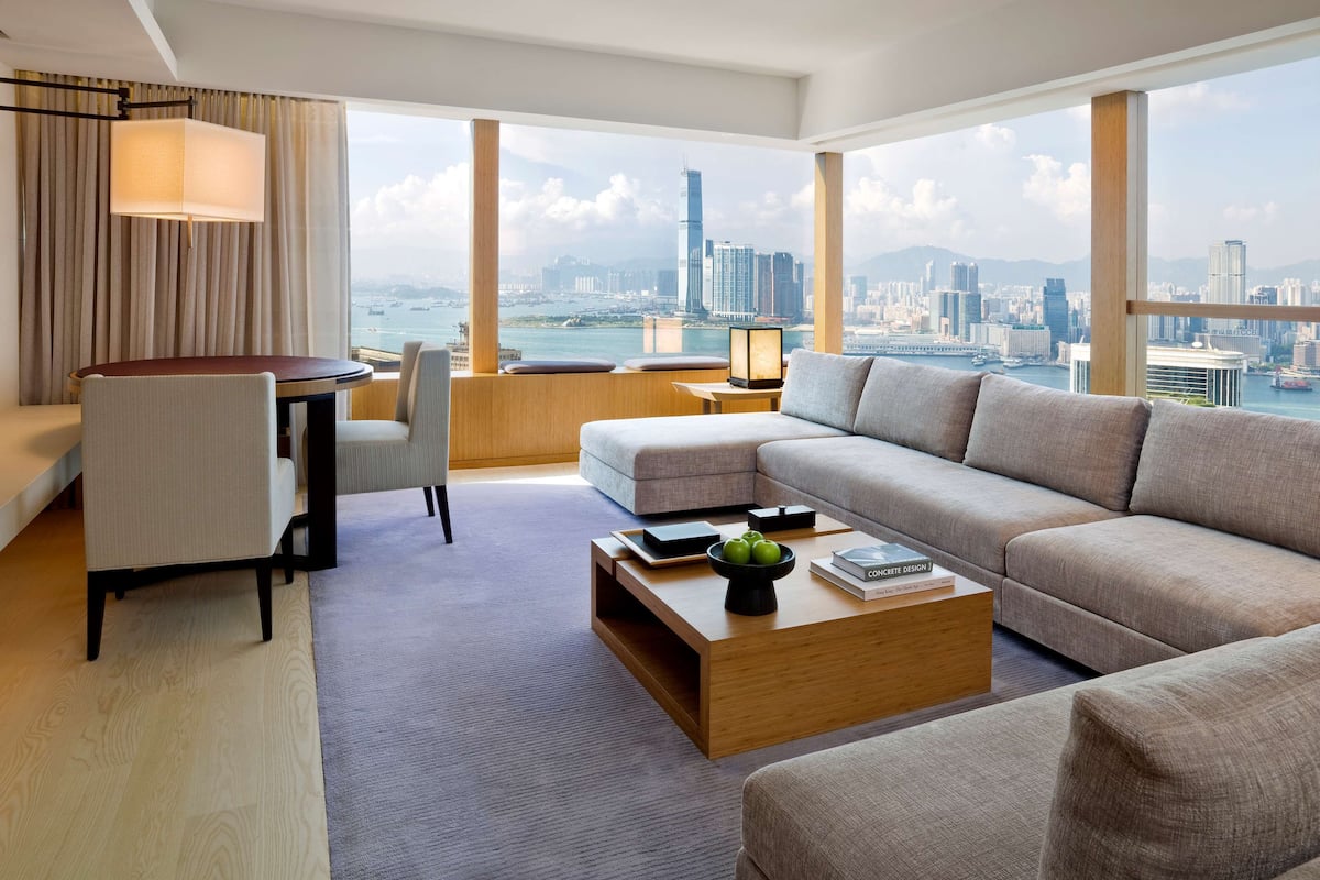 A modern hotel room with a large window offering a city view, a sofa, a coffee table, and a desk with a lamp.